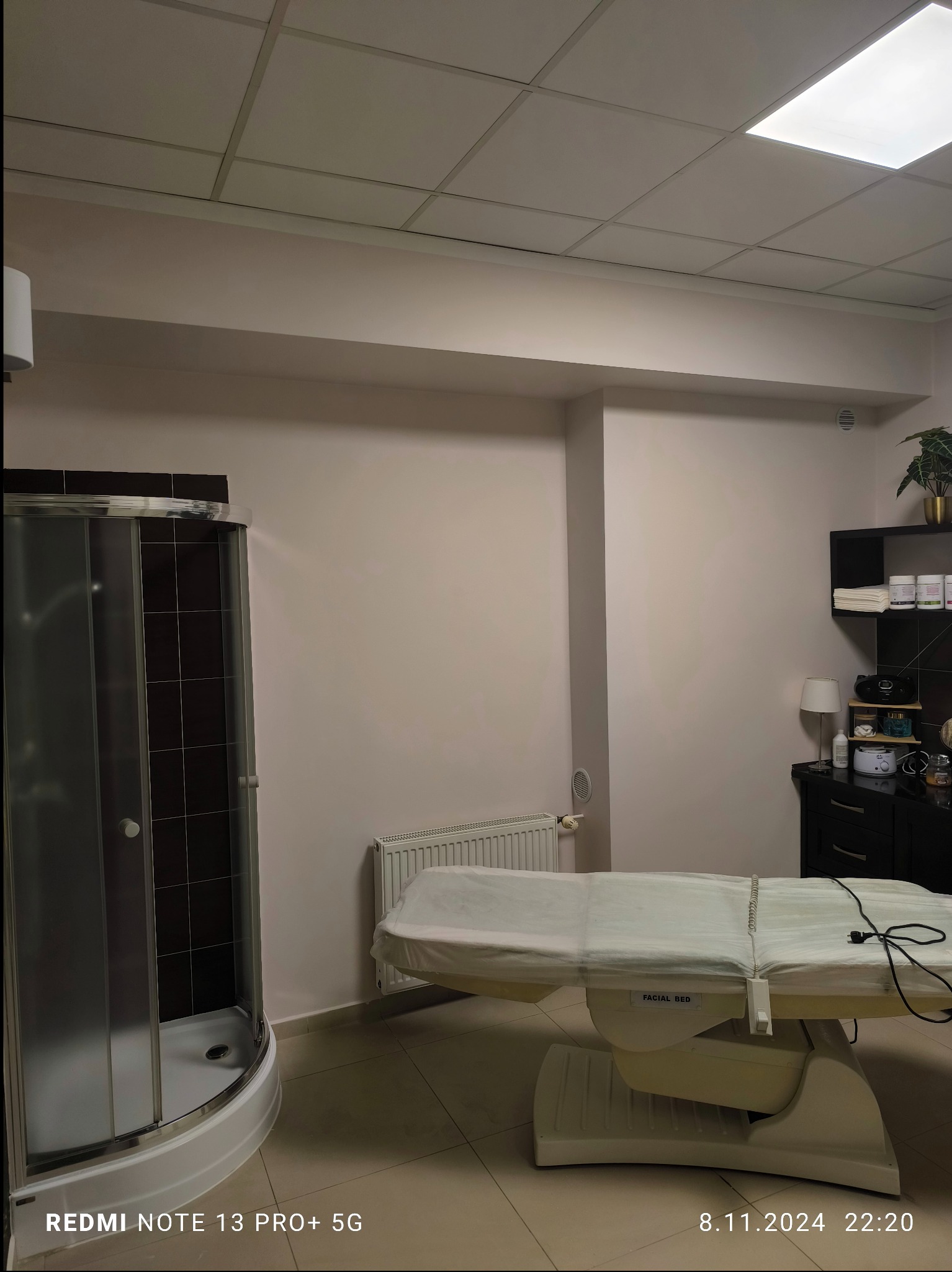 Wnętrze gabinetu kosmetycznego z łóżkiem do zabiegów, kabiną prysznicową i czarną komodą z dekoracjami.