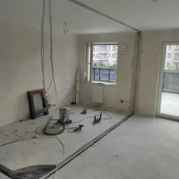 Wnętrze w trakcie demontażu: widoczne bruzdy w podłodze, kable zwisające z sufitu, odkurzacz przemysłowy, poziomica i narzędzia na podłodze, wiadro oraz fragment wyciętej podłogi.