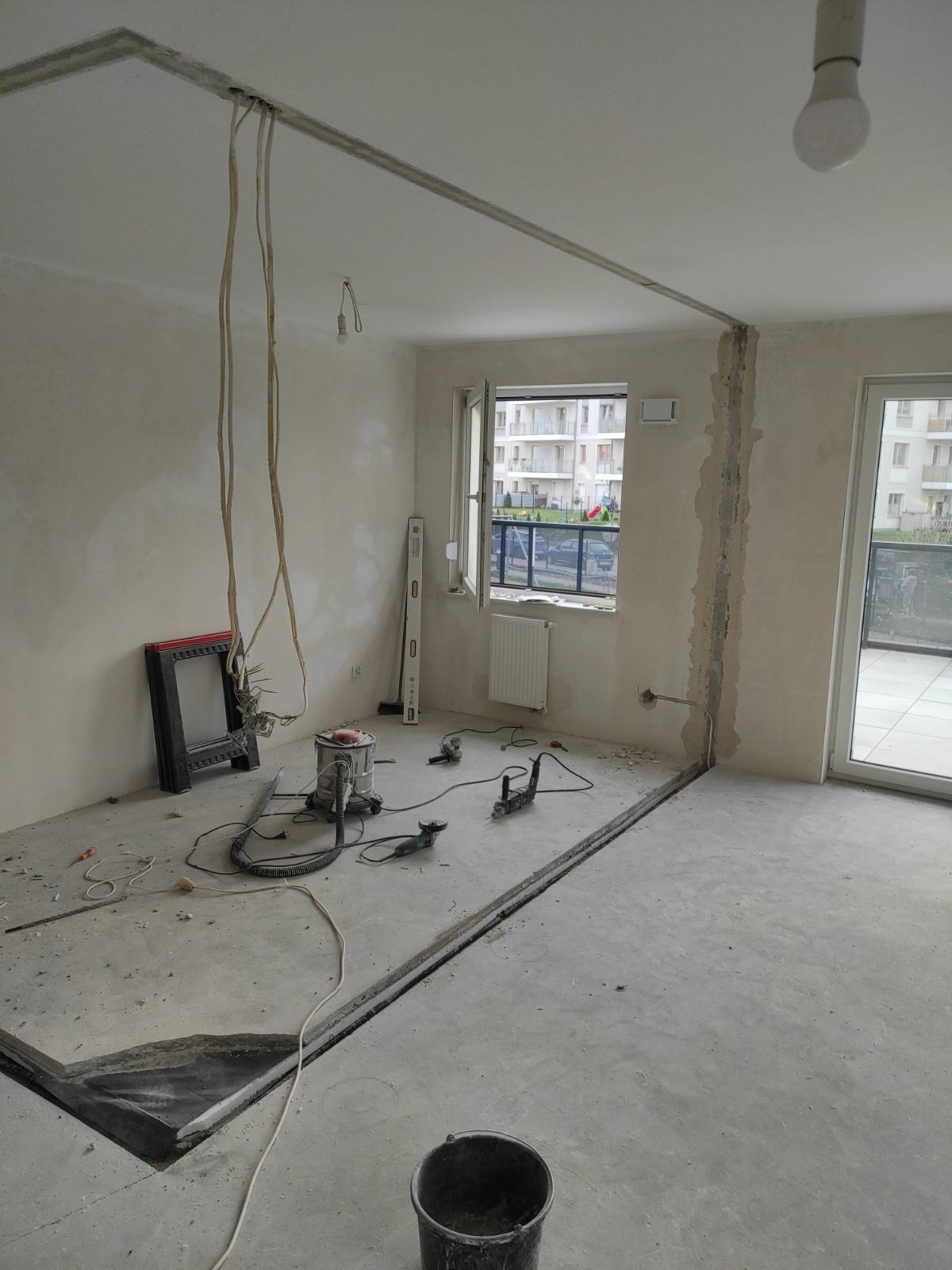 Wnętrze w trakcie demontażu: widoczne bruzdy w podłodze, kable zwisające z sufitu, odkurzacz przemysłowy, poziomica i narzędzia na podłodze, wiadro oraz fragment wyciętej podłogi.
