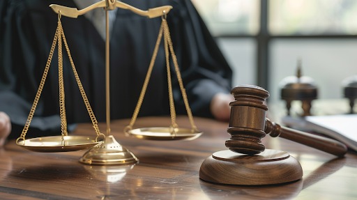 Złota waga sprawiedliwości i drewniany młotek sędziowski na biurku z ciemnego drewna, w tle postać w todze.