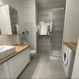 Nowoczesna łazienka z białymi szafkami pod umywalką z drewnianym blatem, kabiną prysznicową z wzorzystymi płytkami i pralką pod blatem.