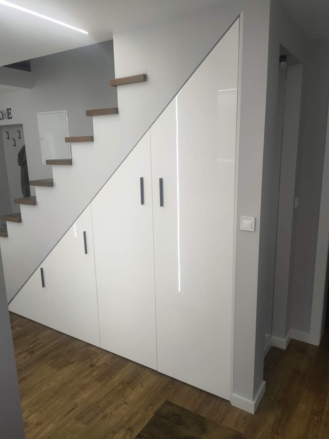 Biała szafa wnękowa pod schodami z drewnianymi stopniami, minimalistyczne czarne uchwyty, panele podłogowe w odcieniach brązu.