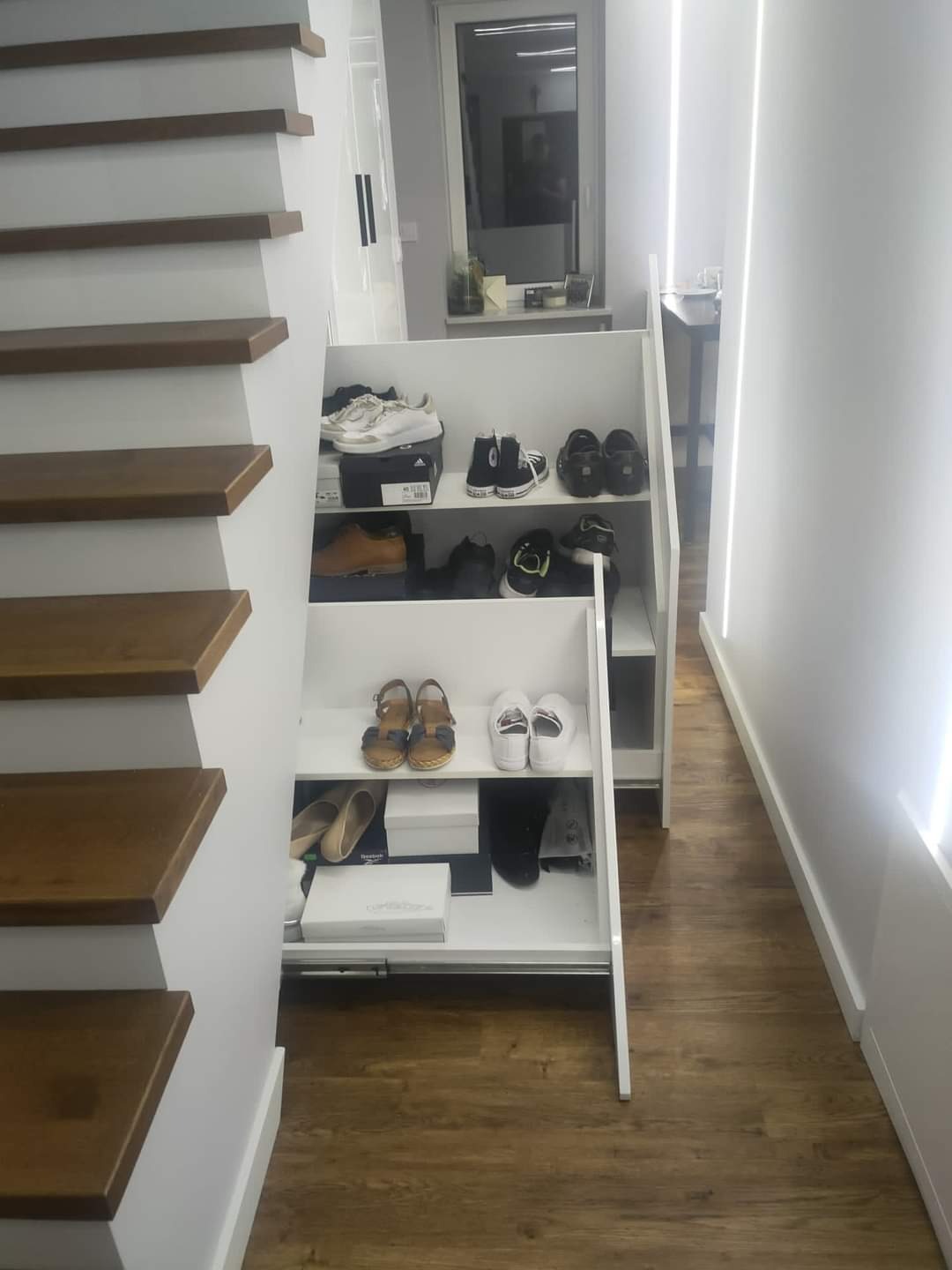 Biała szafka na buty wbudowana pod schodami, z widoczną kolekcją obuwia na półkach i w szufladzie, na tle drewnianej podłogi i minimalistycznego korytarza.