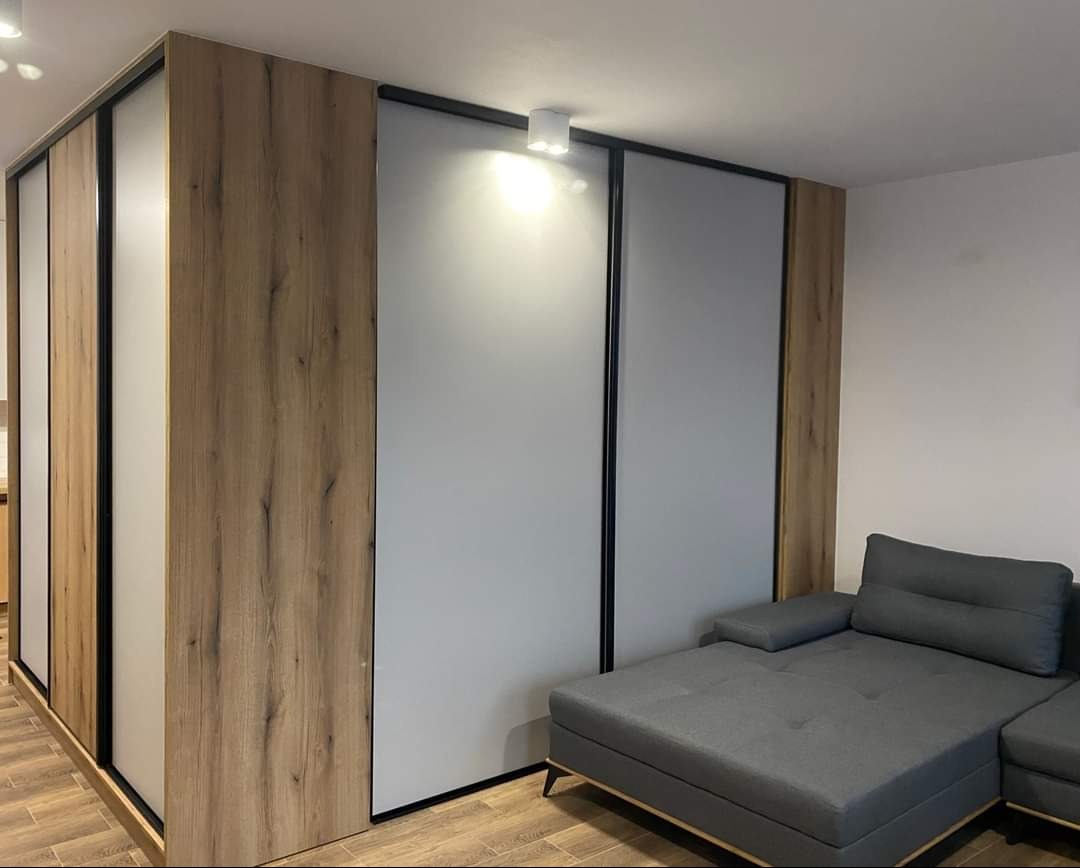 Nowoczesna szafa przesuwna z drewnopodobnymi panelami i szarymi drzwiami, czarne obramowania, widoczna część szarej sofy w minimalistycznym wnętrzu.