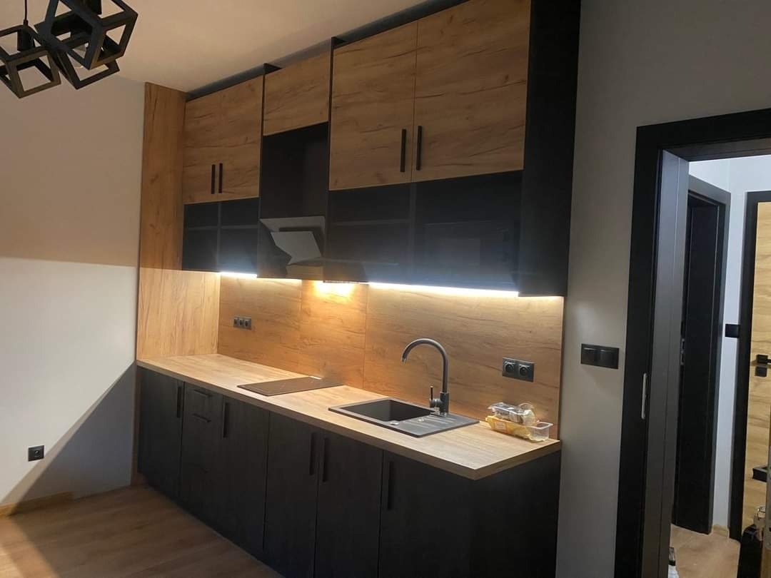 Nowoczesna kuchnia na wymiar z czarnymi szafkami dolnymi i górnymi w odcieniach drewna, podświetlona listwą LED pod szafkami wiszącymi, czarny zlew i bateria, widoczny fragment sufitu z lampą.