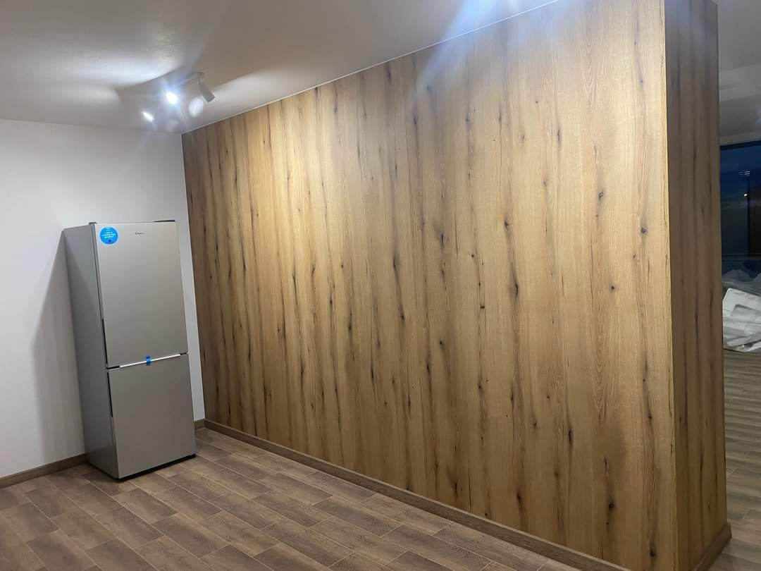 Wnętrze z drewnopodobną zabudową ściany, szara lodówka przy białej ścianie, podłoga z paneli imitujących drewno, oświetlenie punktowe.