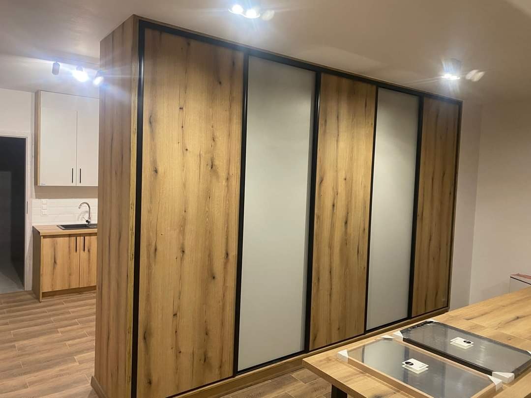 Duża szafa przesuwna z drewnopodobnymi panelami i szarymi wstawkami, czarne obramowanie, widoczna kuchnia w tle z drewnianymi szafkami i białymi górnymi szafkami.