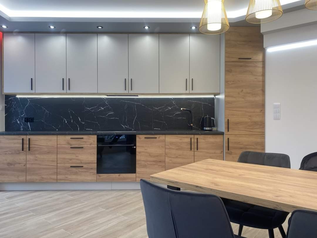 Nowoczesna kuchnia z szarymi górnymi szafkami i drewnopodobnymi dolnymi szafkami, czarny blat z marmurowym wzorem, oświetlenie LED pod szafkami, drewniany stół z szarymi krzesłami w tle.