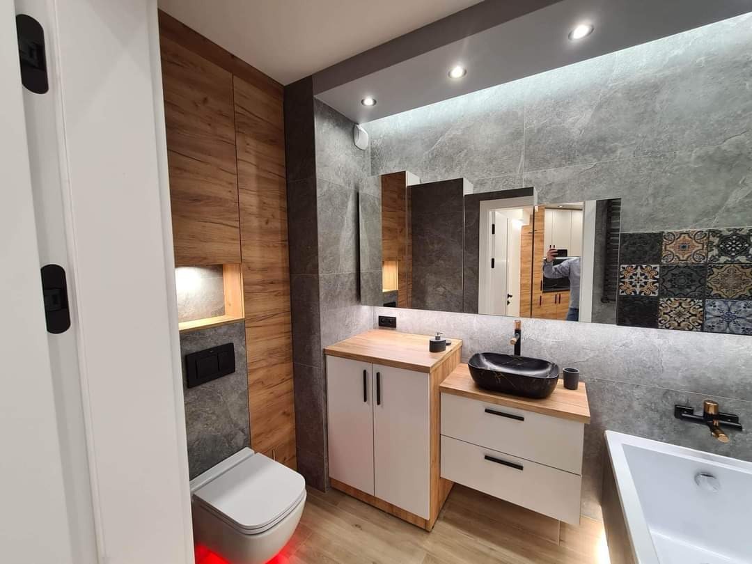 Nowoczesna łazienka z drewnianymi akcentami, szarą glazurą imitującą kamień, czarną umywalką nablatową i toaletą podświetloną na czerwono.