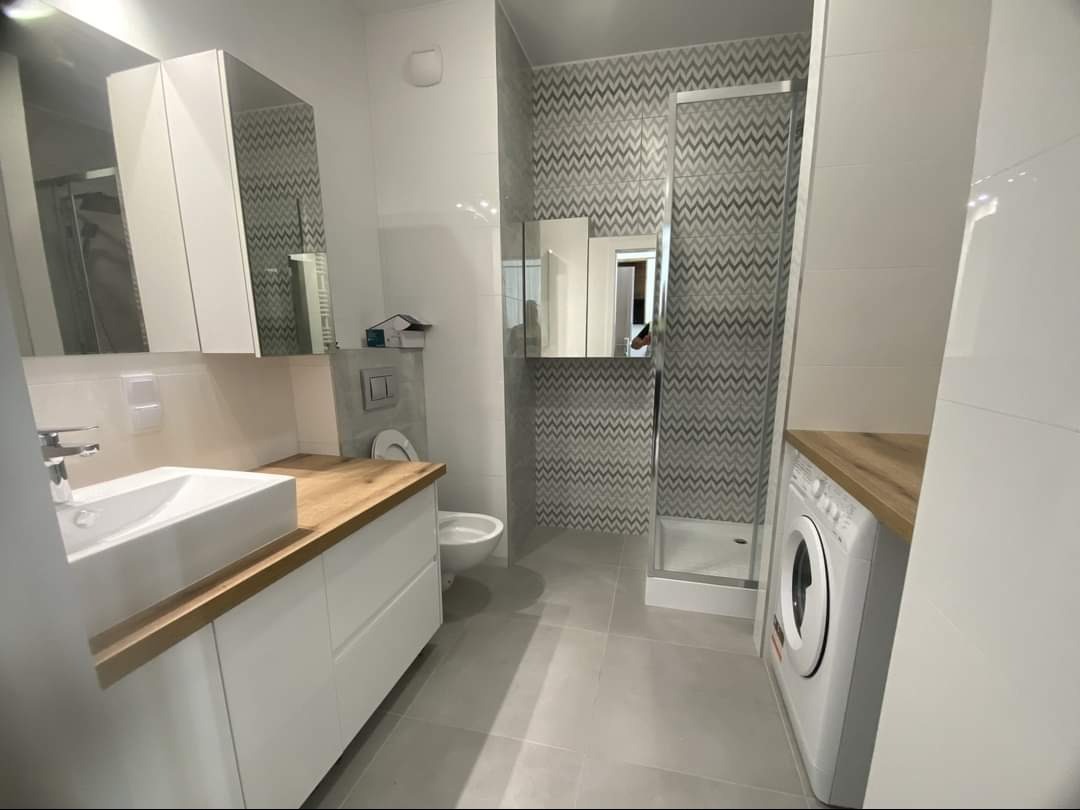 Nowoczesna łazienka z białymi szafkami pod umywalką z drewnianym blatem, kabiną prysznicową z wzorzystymi płytkami i pralką pod blatem.