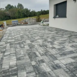 Wykończony podjazd z szarej kostki brukowej ułożonej wzorem. W tle, przy białej ścianie budynku, leżą zapasy kostki. Prace brukarskie w toku, widoczny pracownik.