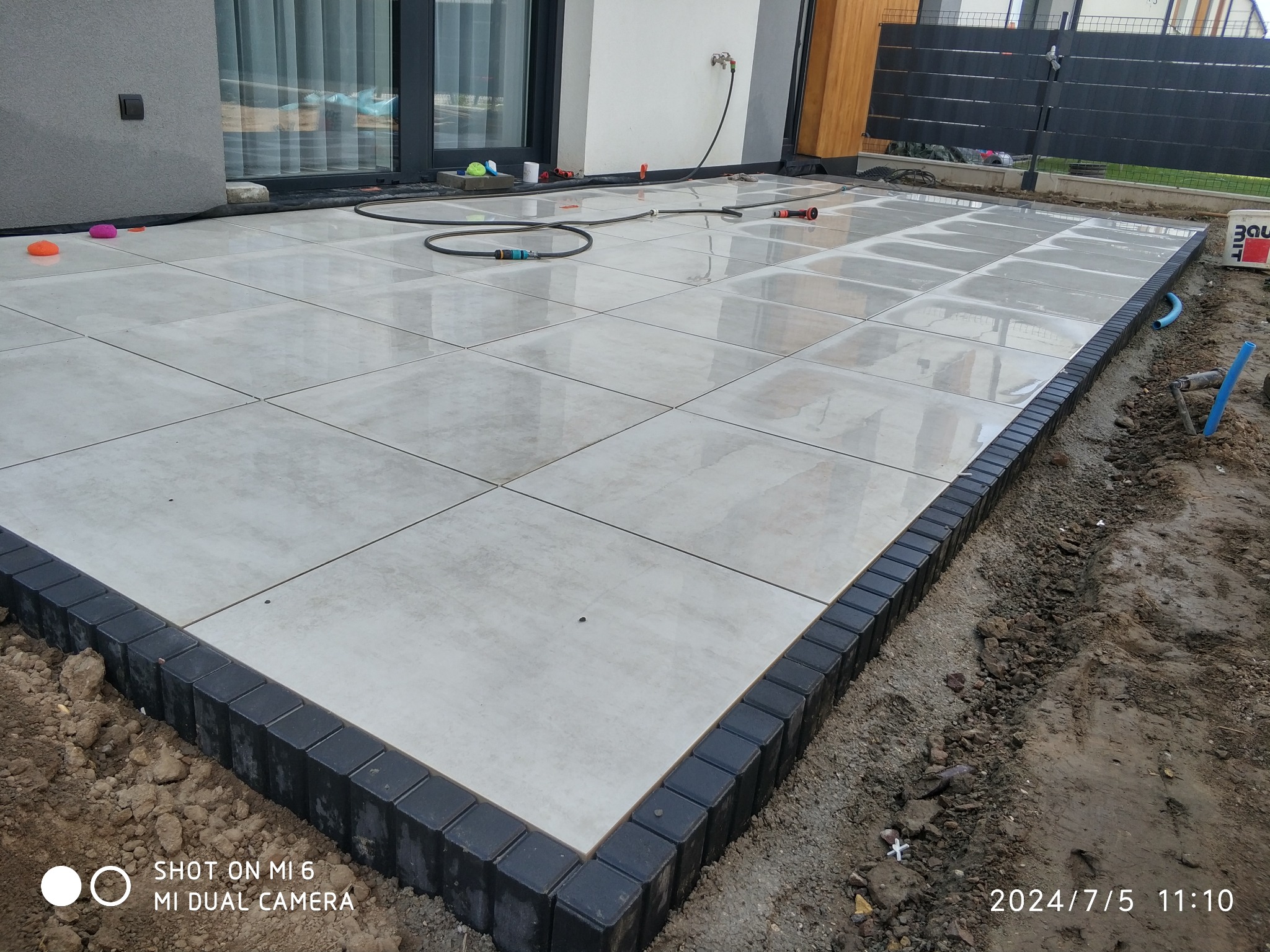 Jasne płytki tarasowe z ciemną obwódką, otoczone ziemią, z widocznym wężem ogrodowym i zabawkami, sugerujące świeżo ukończone prace brukarskie.