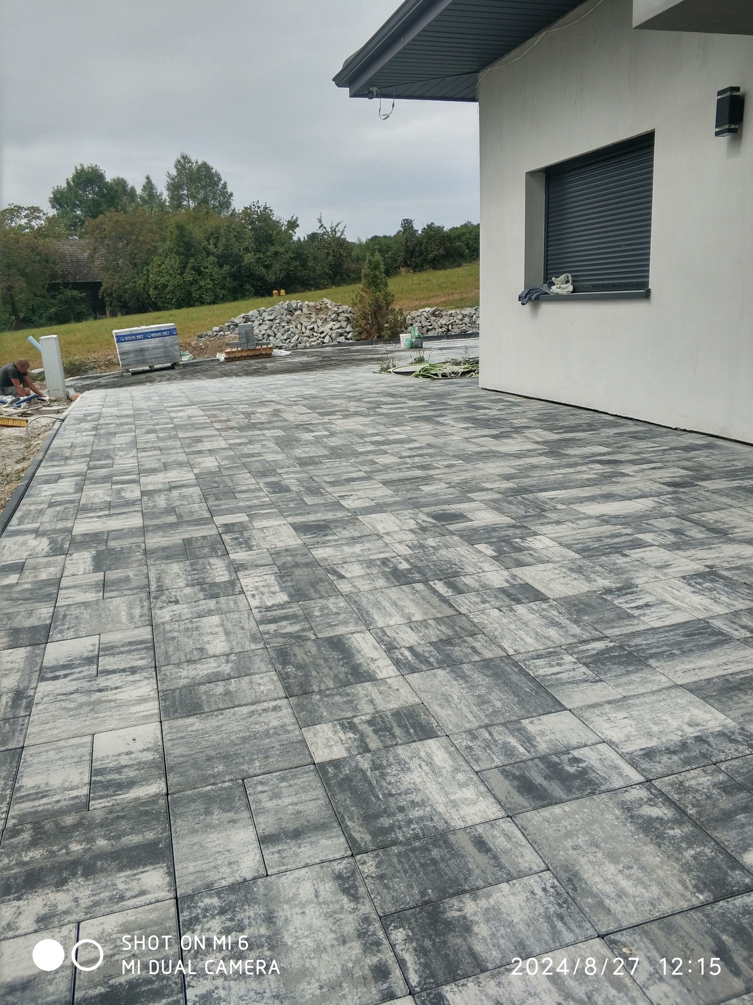 Wykończony podjazd z szarej kostki brukowej ułożonej wzorem. W tle, przy białej ścianie budynku, leżą zapasy kostki. Prace brukarskie w toku, widoczny pracownik.