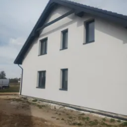 Elewacja domu jednorodzinnego z gładzią, widoczne okna z czarnymi ramami, dach z ciemną dachówką i rynną, na podwórku stoi biały bus.