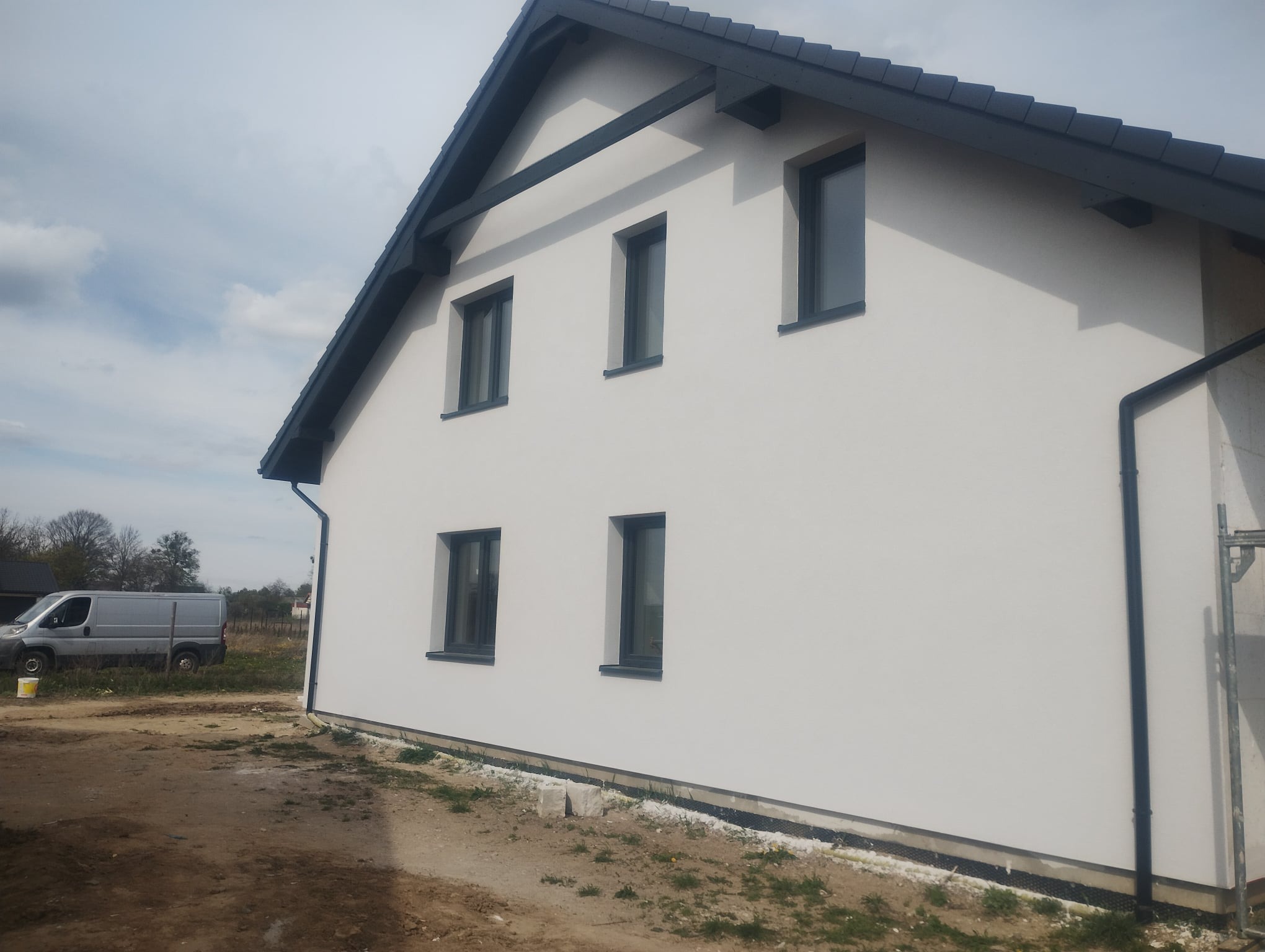 Elewacja domu jednorodzinnego z gładzią, widoczne okna z czarnymi ramami, dach z ciemną dachówką i rynną, na podwórku stoi biały bus.