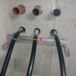 PROFI-ELEKTRO Usługi instalatorsko-eletryczne MARCIN ZIOBROŃ - Betonowa ściana z trzema rurami instalacyjnymi i trzema grubymi kablami elektrycznymi zamocowanymi na metalowej szynie.