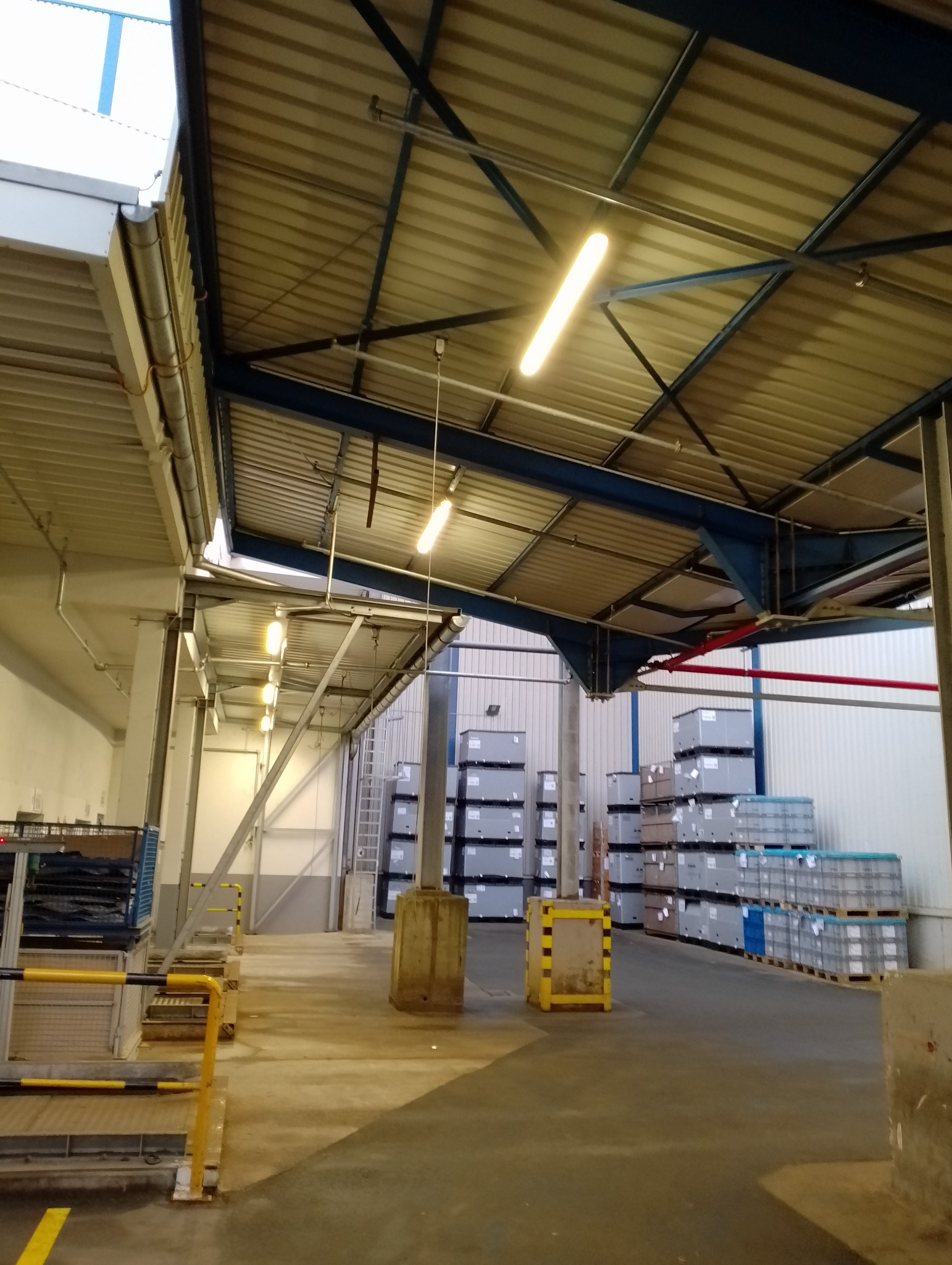 Wnętrze hali magazynowej z rzędami metalowych regałów i palet z szarymi pojemnikami, oświetlonej podłużnymi lampami jarzeniowymi pod metalowym dachem z widoczną konstrukcją stalową.