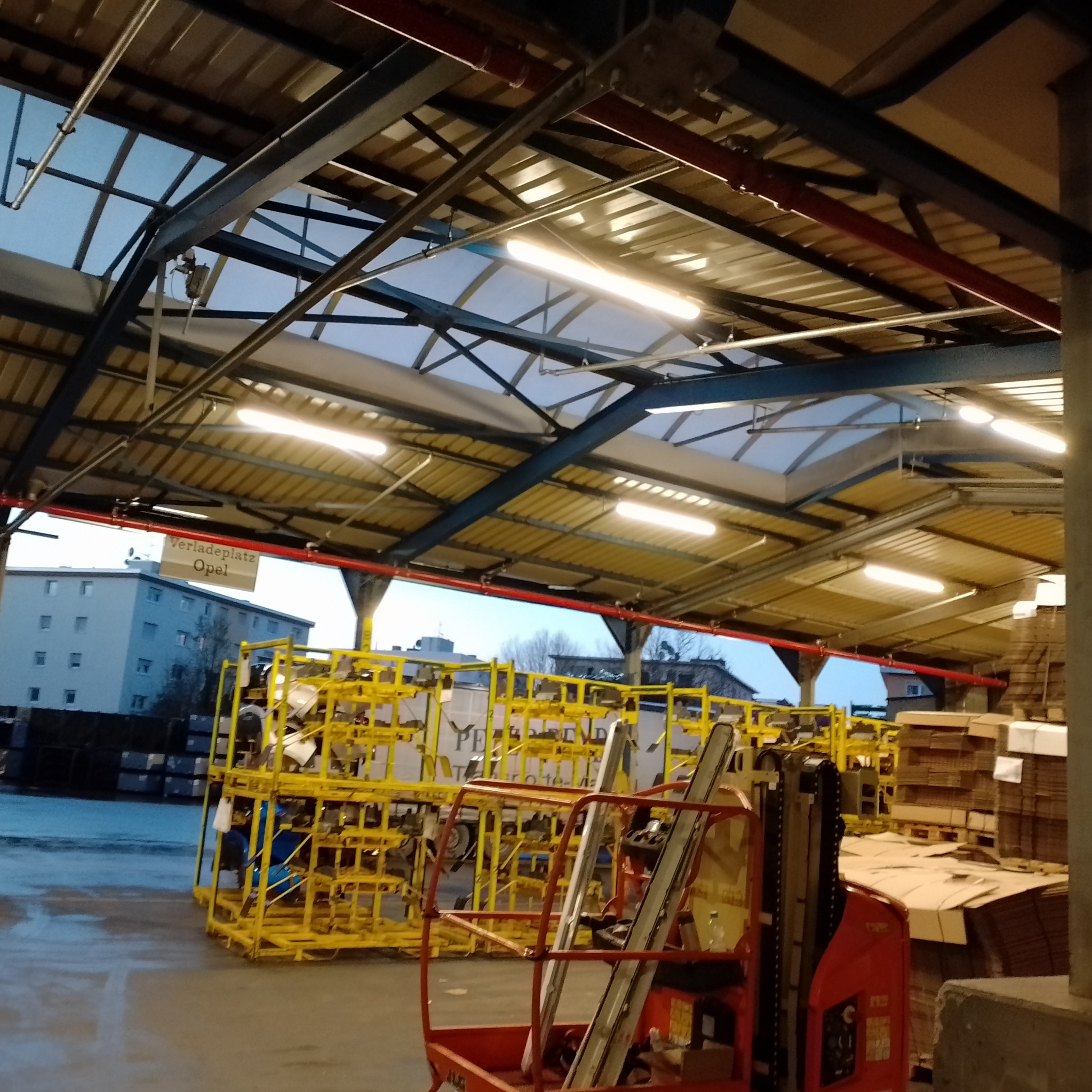 Wnętrze hali magazynowej z widocznymi lampami jarzeniowymi pod sufitem, żółtymi konstrukcjami i podnośnikiem koszowym.