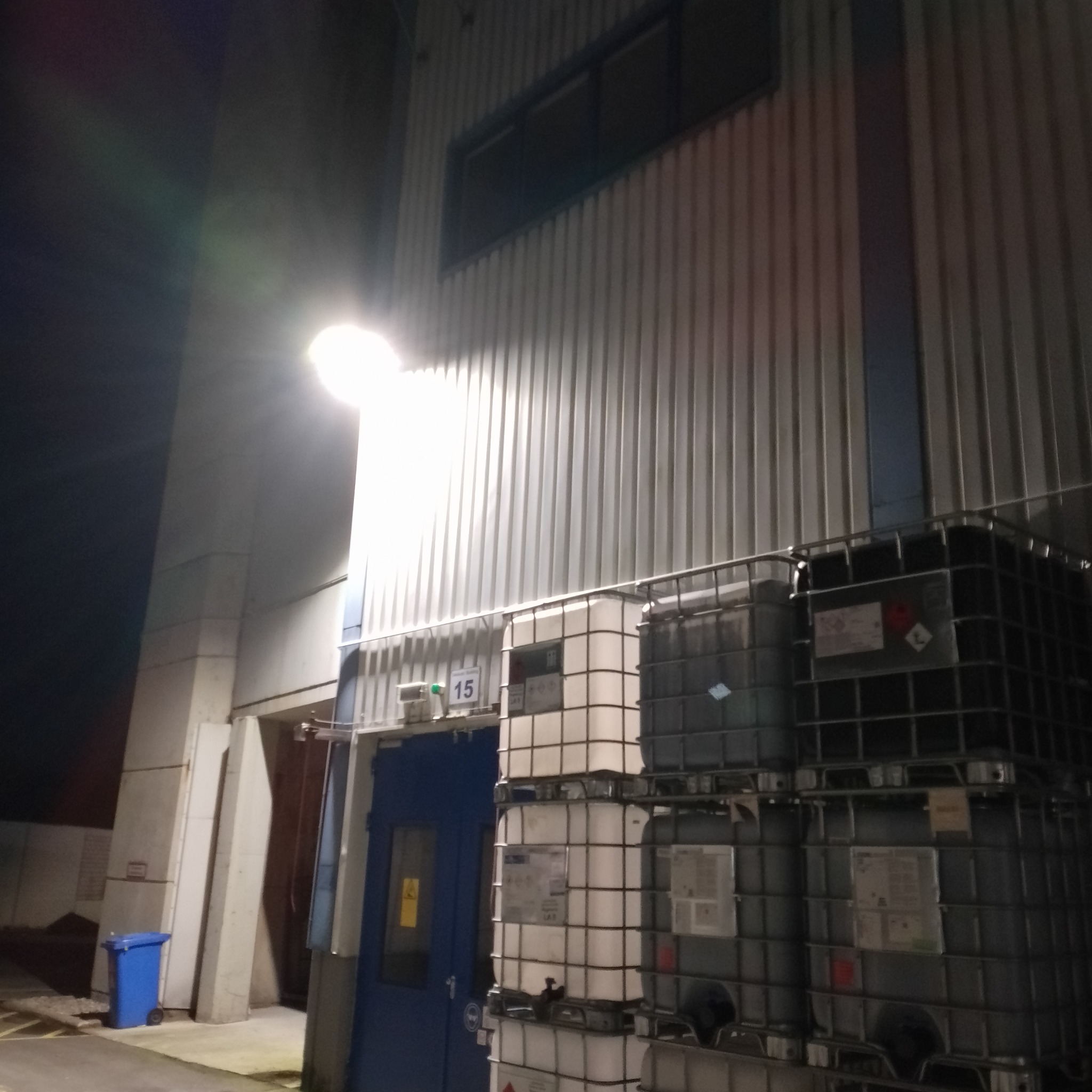 Zewnętrzne oświetlenie przemysłowe budynku z blachy trapezowej, z widocznymi zbiornikami IBC i niebieskim koszem na śmieci