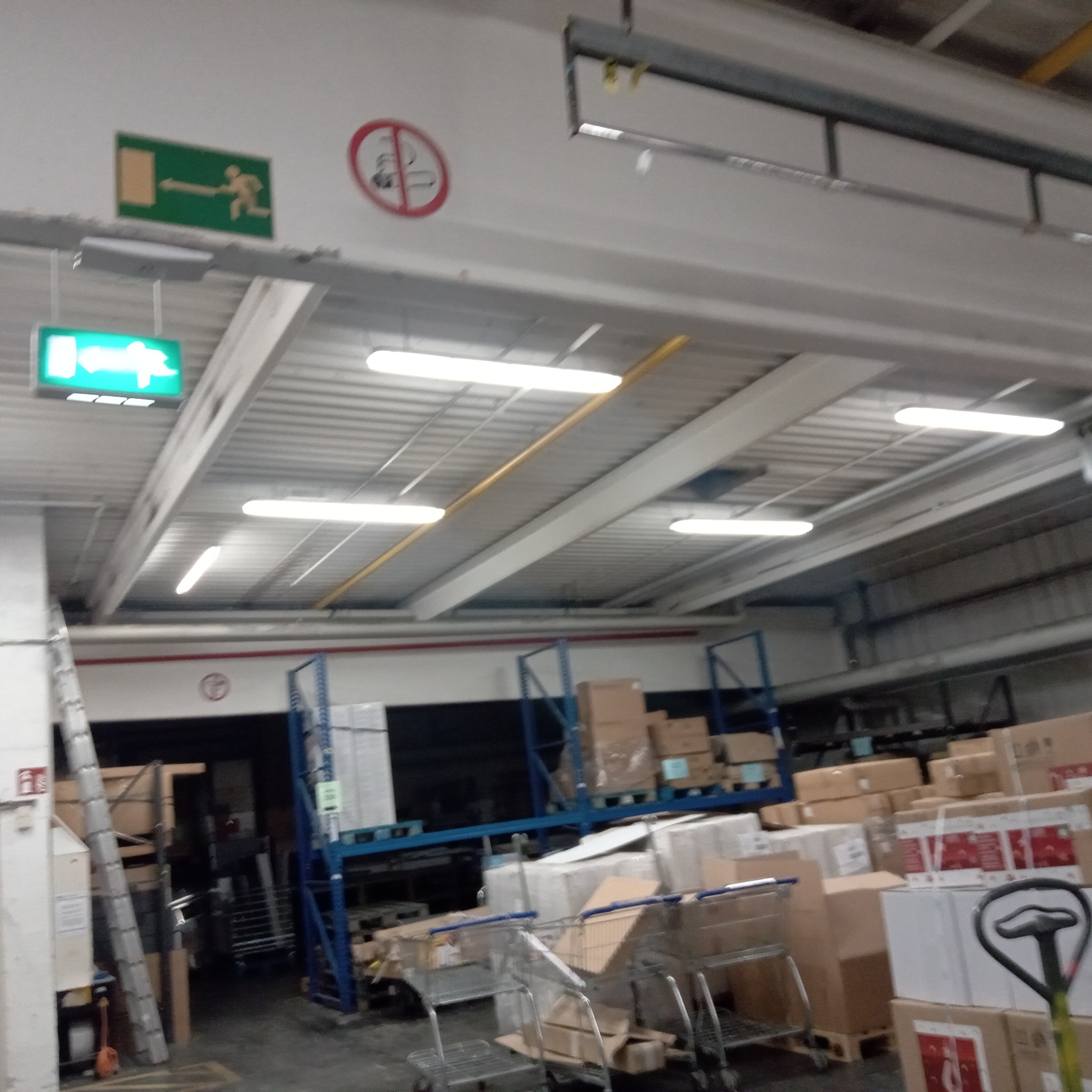 Wnętrze magazynu z oświetleniem LED, widoczne znaki ewakuacyjne i zakazu palenia, regały z kartonami i wózki transportowe.