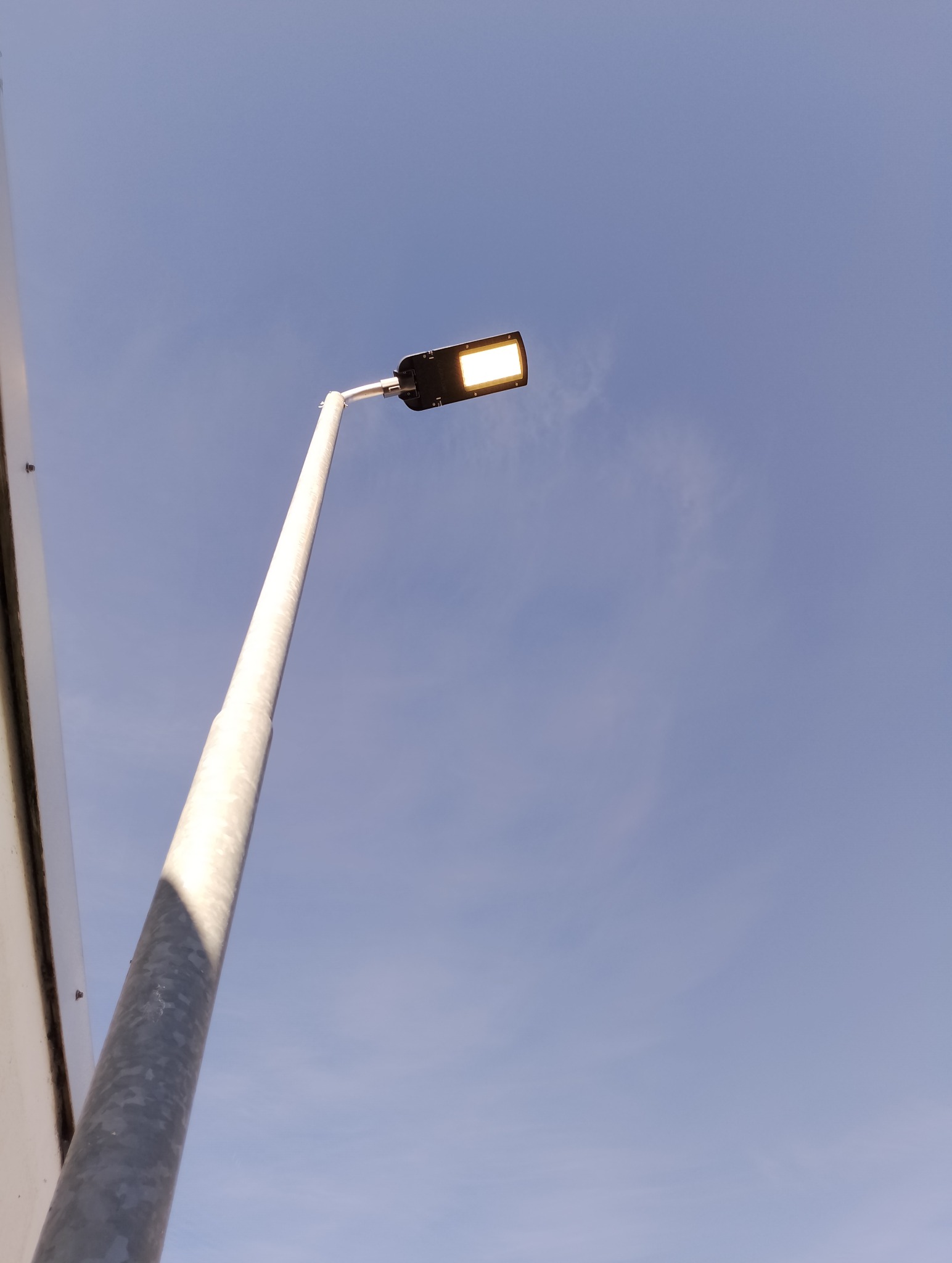 Pojedyncza, nowoczesna lampa uliczna LED z metalowym słupem oświetlona od dołu, na tle błękitnego nieba.