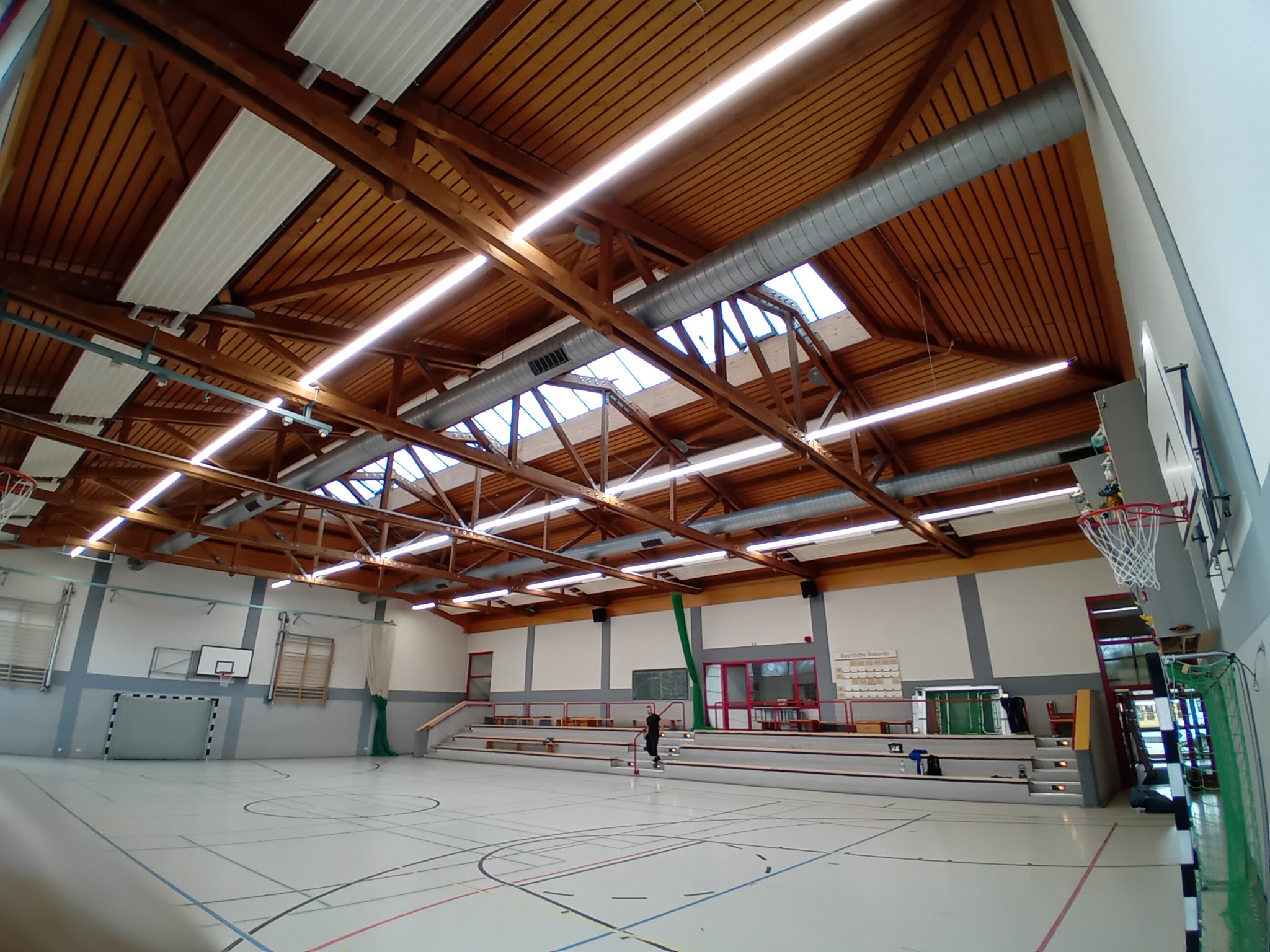 Wnętrze hali sportowej z drewnianą konstrukcją dachu, widocznymi instalacjami wentylacyjnymi i oświetleniem liniowym LED.