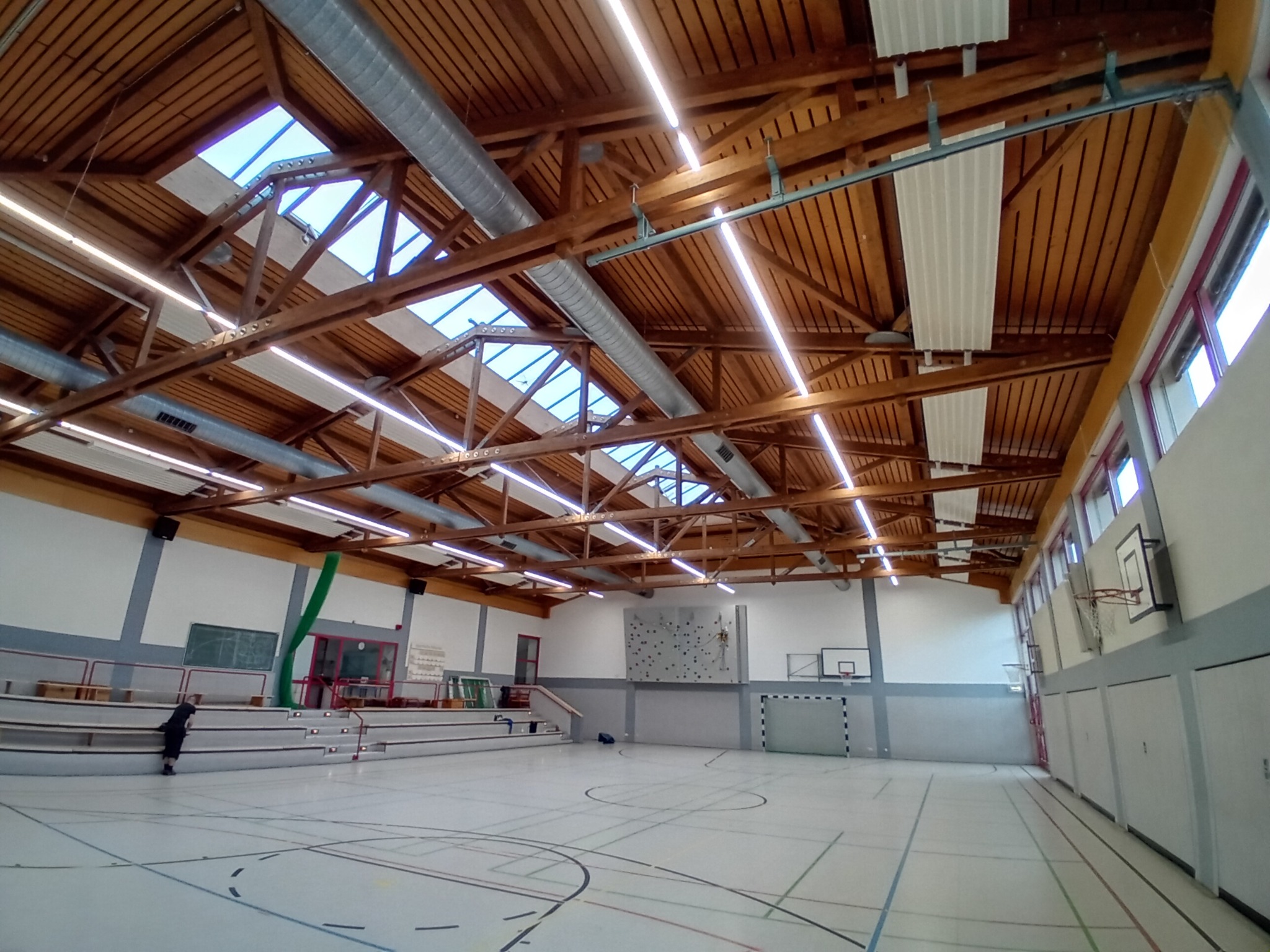 Wnętrze hali sportowej z drewnianą konstrukcją dachu, oświetleniem liniowym LED, widocznymi instalacjami wentylacyjnymi i świetlikami dachowymi.