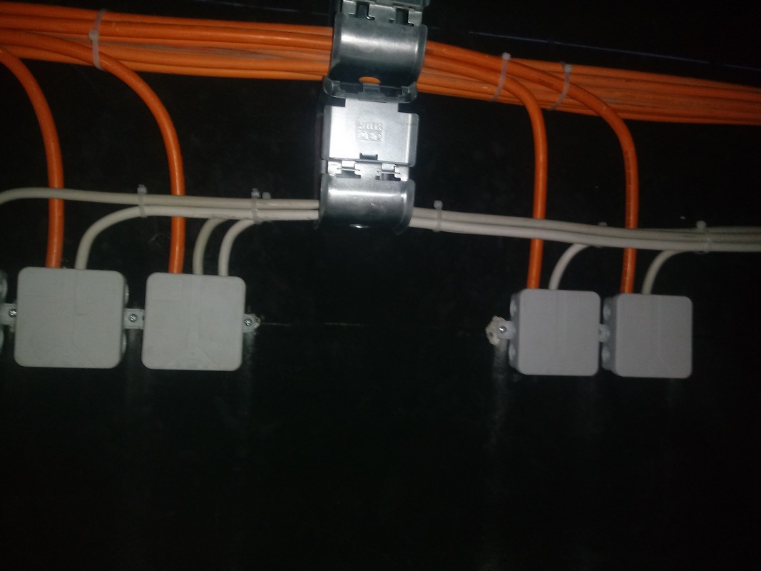 Szereg białych puszek instalacyjnych na czarnej ścianie, połączonych białymi i pomarańczowymi kablami, mocowanych metalowymi uchwytami.