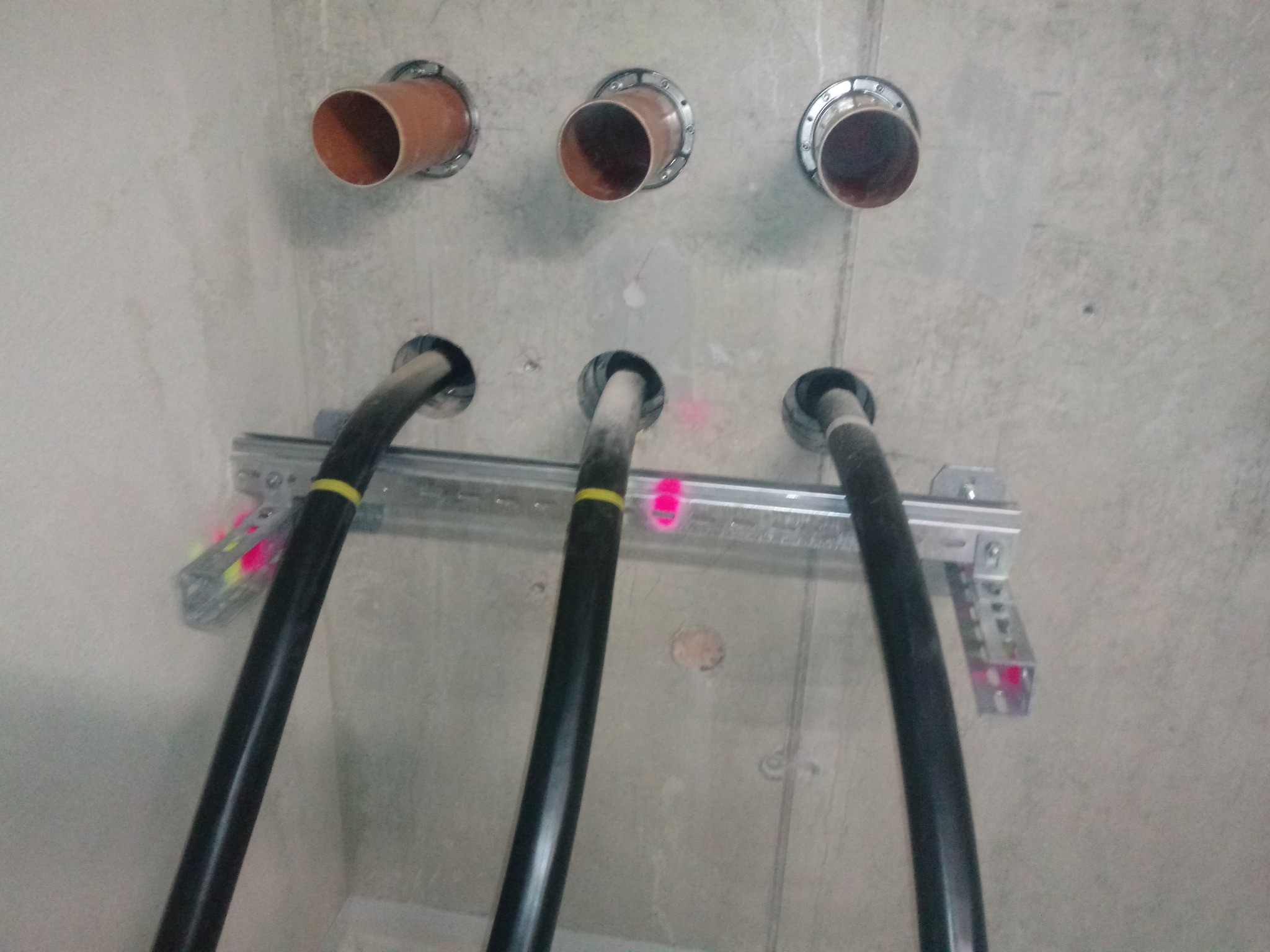 Betonowa ściana z trzema rurami instalacyjnymi i trzema grubymi kablami elektrycznymi zamocowanymi na metalowej szynie.
