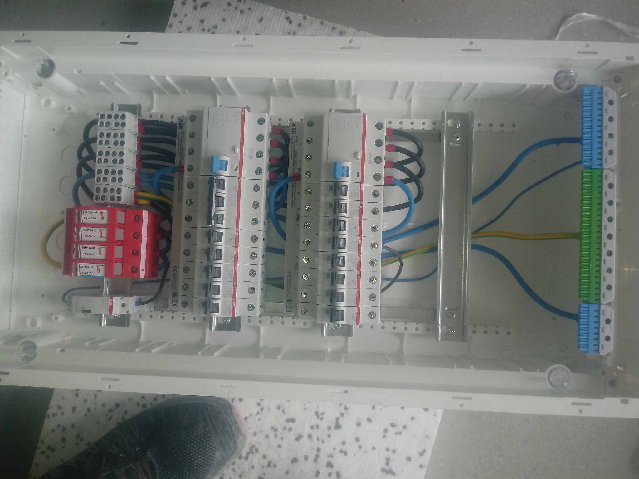 Rozdzielnica elektryczna w białej obudowie z widocznymi zabezpieczeniami przepięciowymi DEHNguard, bezpiecznikami ABB, listwami zaciskowymi w kolorach niebieskim i zielonym oraz okablowaniem...