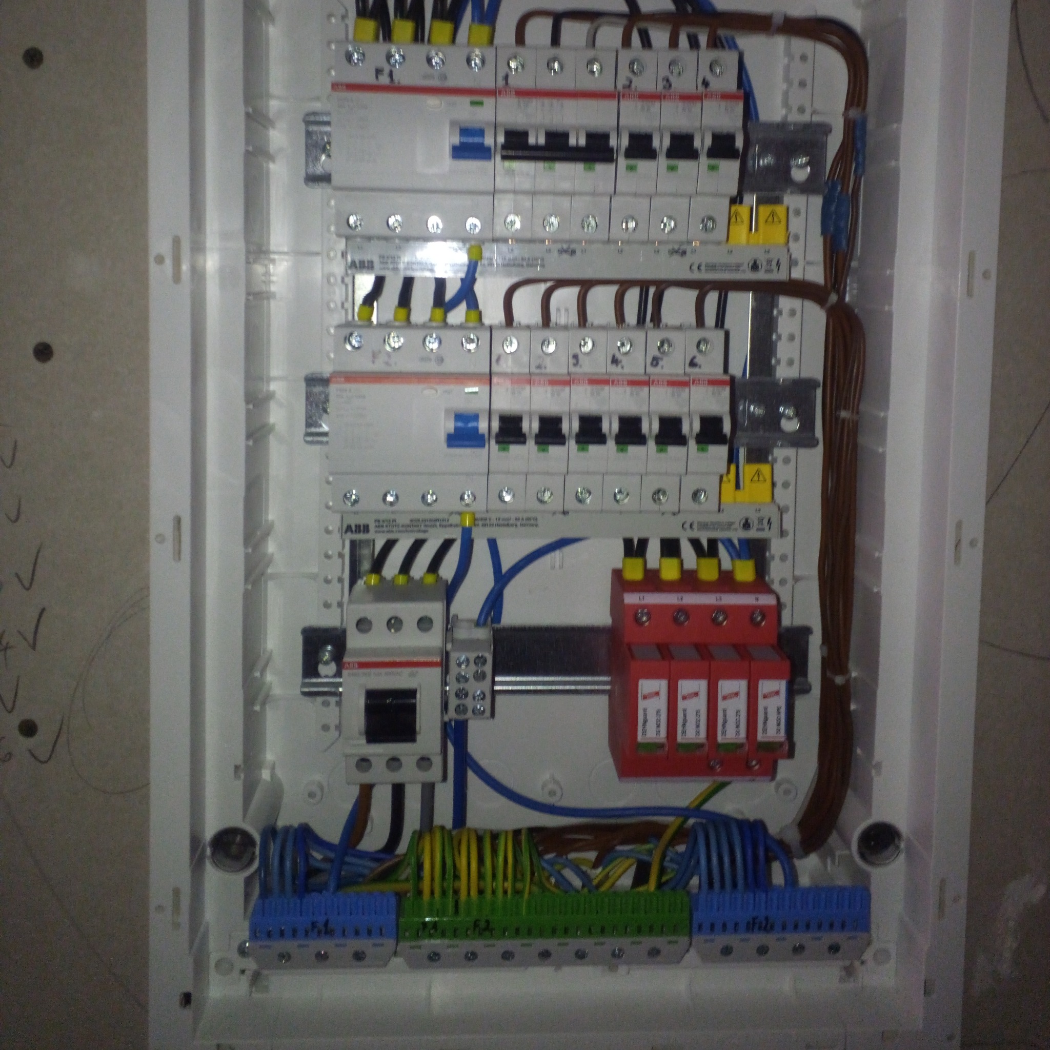 Rozdzielnica elektryczna w białej obudowie z widocznymi bezpiecznikami, przewodami w różnych kolorach (niebieski, żółto-zielony, brązowy) oraz ochronnikami przeciwprzepięciowymi, zamontowana...