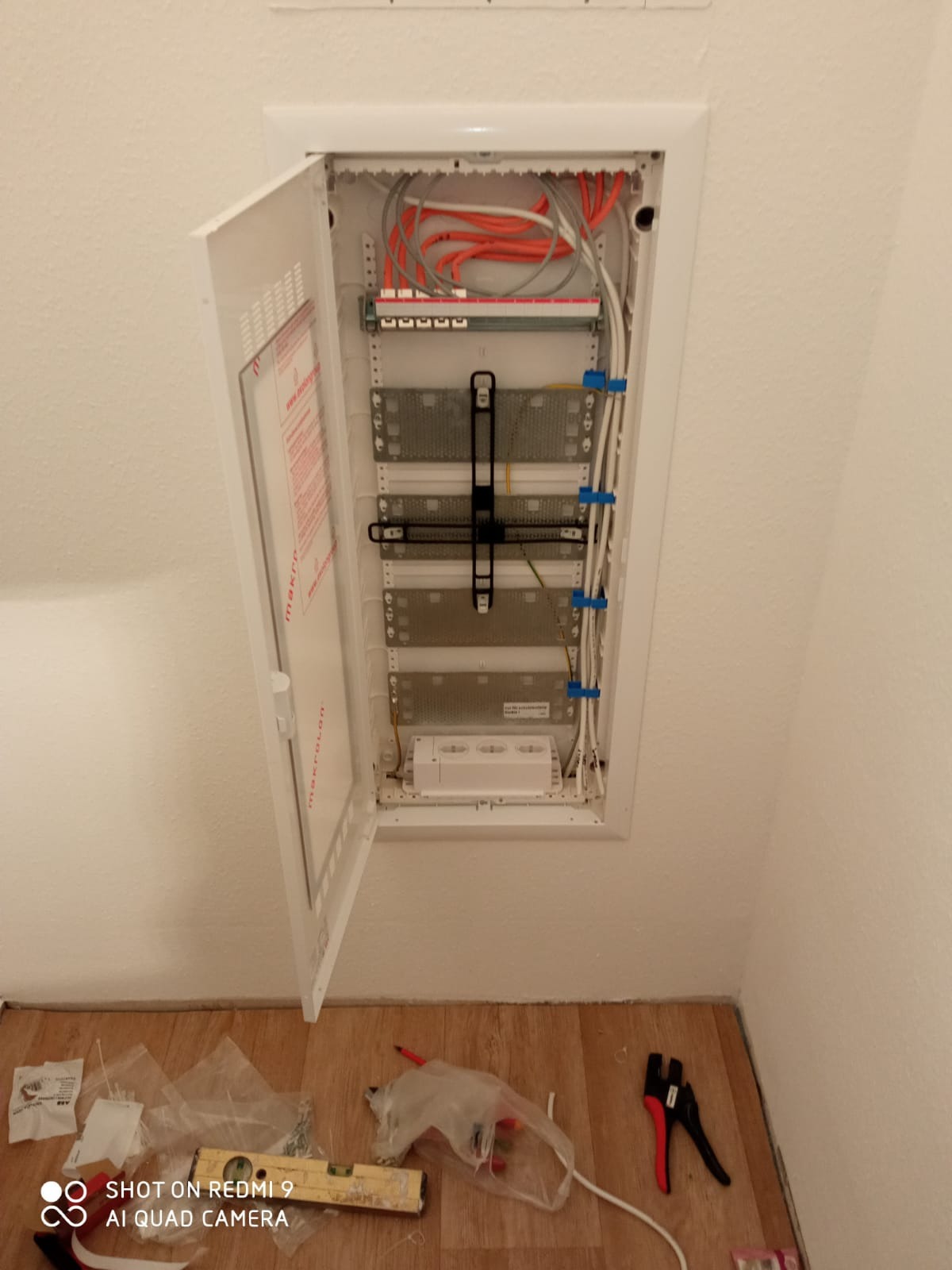 Otwarta szafka elektryczna w ścianie z widocznymi przewodami, listwami zaciskowymi i akcesoriami. Na podłodze leżą narzędzia, poziomica i resztki opakowań po elementach.