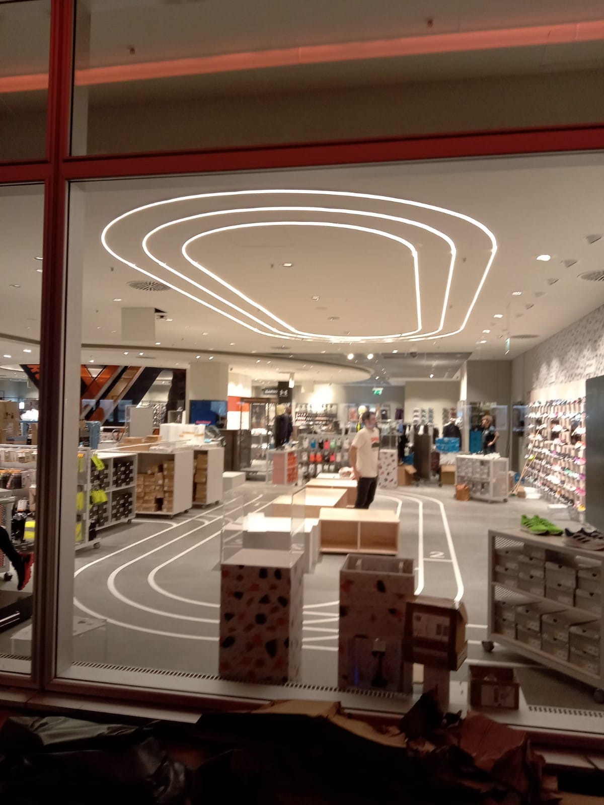 Wnętrze sklepu sportowego z torem biegowym na podłodze i unikalnym oświetleniem sufitowym w kształcie toru, widoczne przez witrynę sklepową.