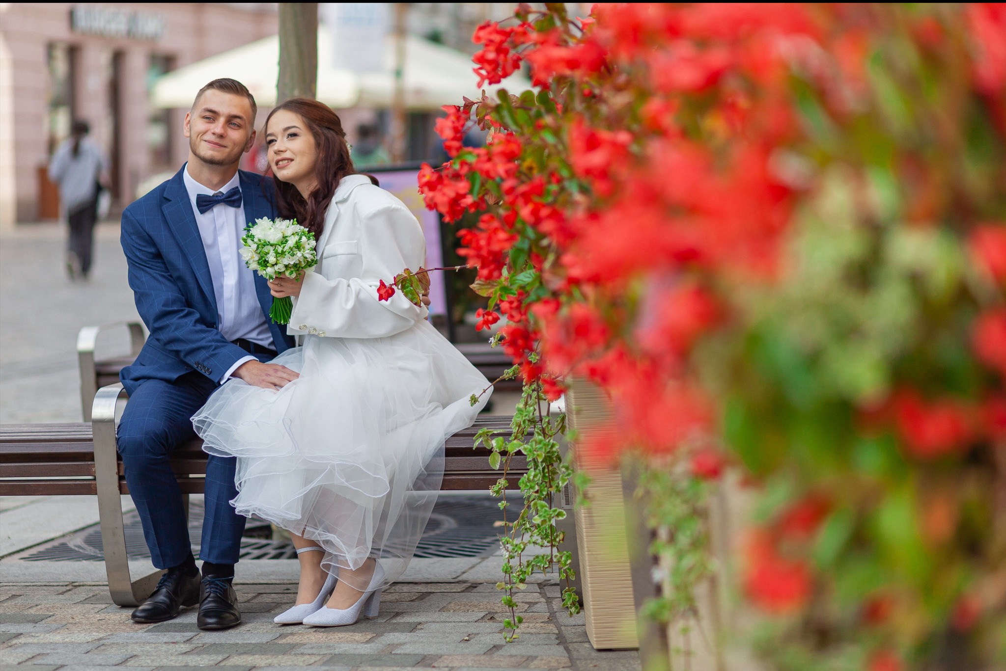 Uśmiechnięta para młoda siedzi na ławce, panna młoda w białej sukni z bukietem, pan młody w niebieskim garniturze z muszką, w tle czerwone kwiaty i budynki.