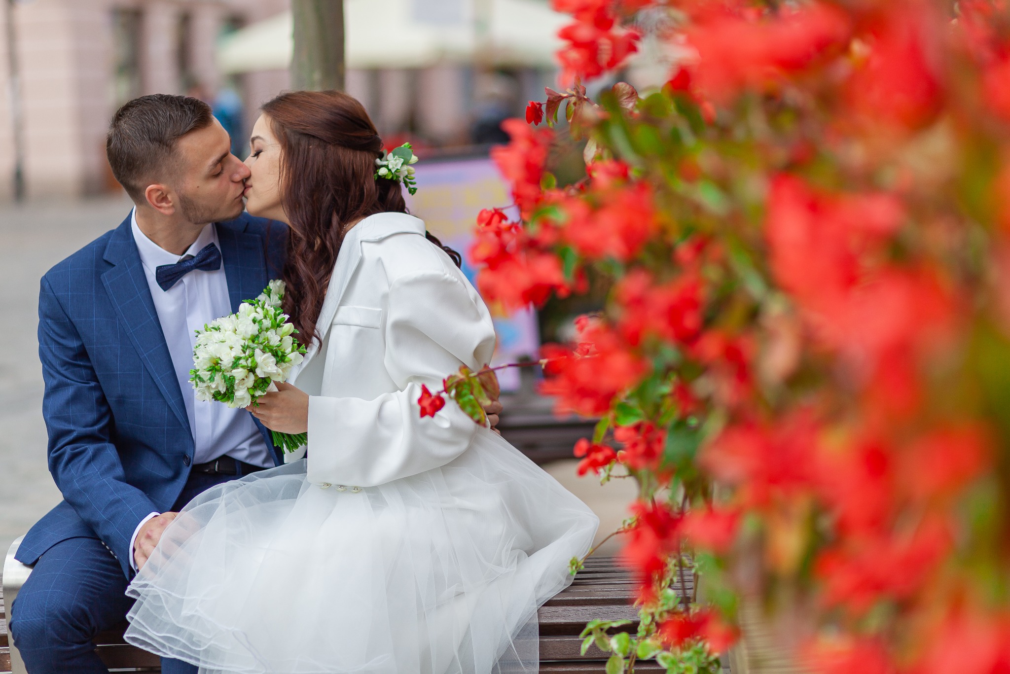 Pocałunek nowożeńców siedzących na ławce, panna młoda trzyma bukiet białych kwiatów, czerwone kwiaty w tle.