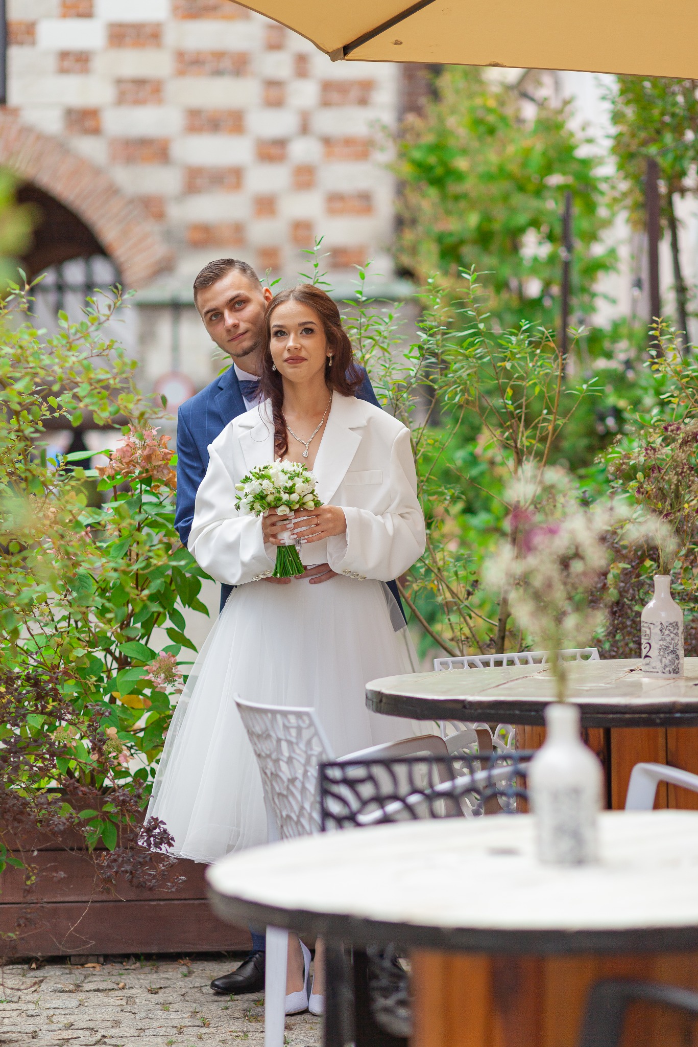 Para młoda w Lublinie pozuje do zdjęcia w plenerze, panna młoda w sukni ślubnej i białym oversize'owym żakiecie trzyma bukiet, pan młody w granatowym garniturze obejmuje ją od tyłu.