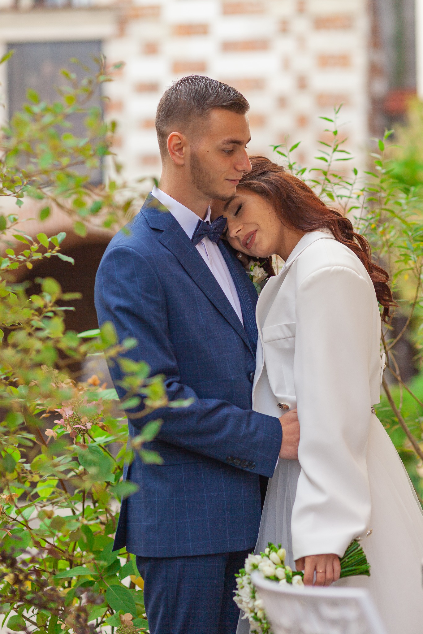 Para młoda w objęciach na tle zieleni: on w granatowym garniturze w kratę i muszce, ona w białej sukni i marynarce, trzyma bukiet.