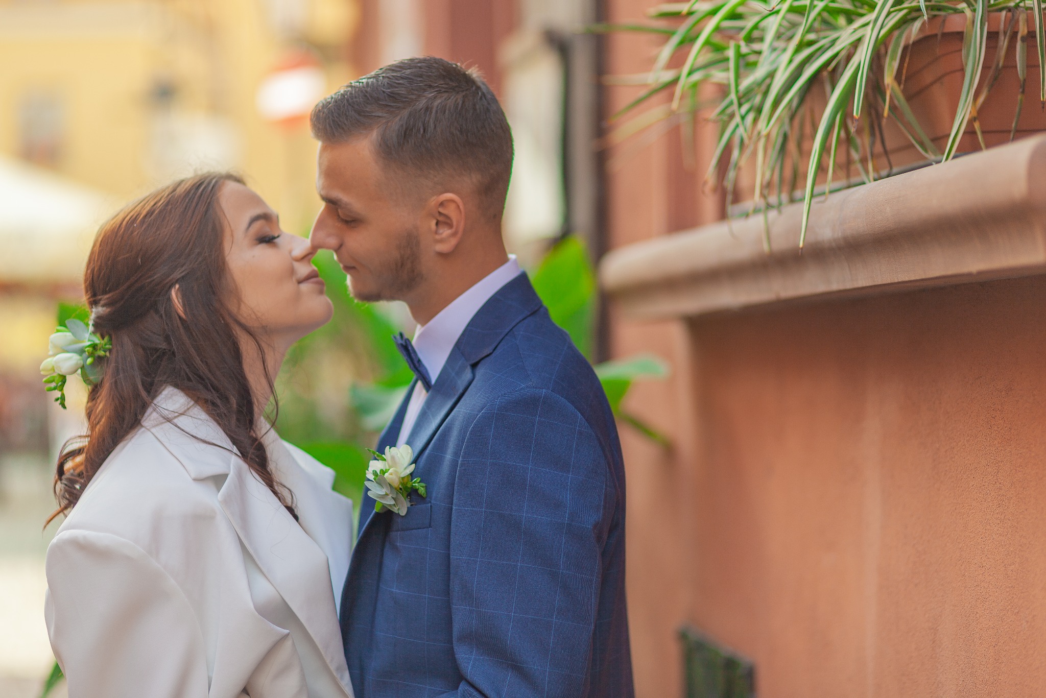 Para młoda w eleganckich strojach, zbliżająca się do pocałunku na tle budynku z roślinami w donicach.