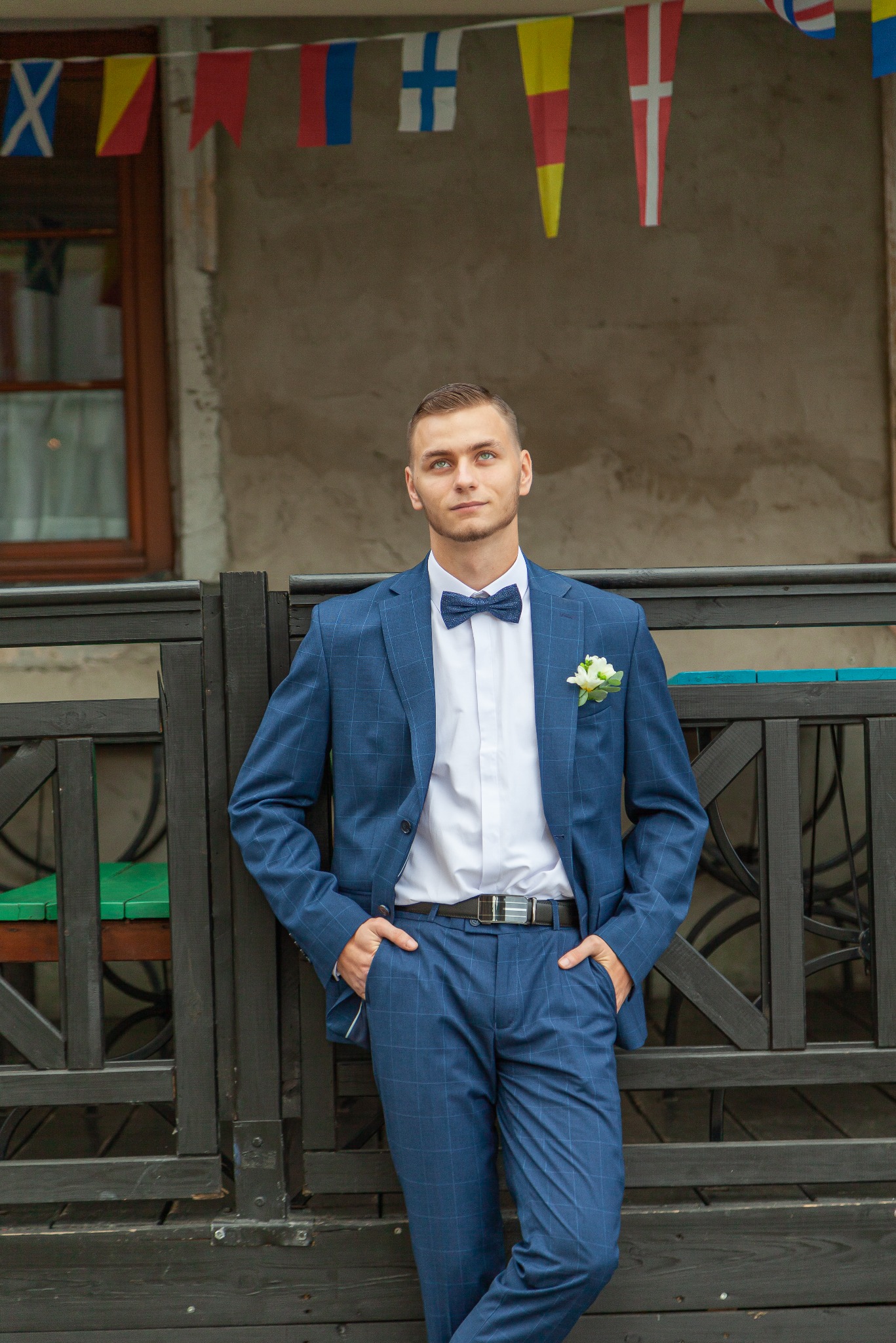 Młody mężczyzna w niebieskim garniturze w kratę i muszce pozuje oparty o czarne drewniane ogrodzenie, z bukiecikiem w butonierce, na tle budynku z flagami.