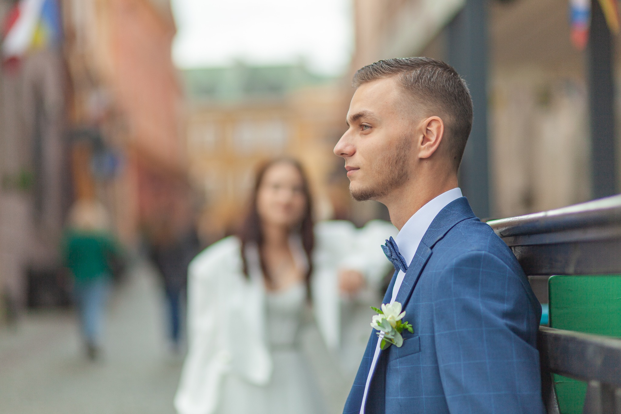 Młody pan młody w niebieskim garniturze w kratę i z muszką, oparty o metalową barierkę, z zamyślonym spojrzeniem, w tle rozmyta panna młoda w białej sukni.