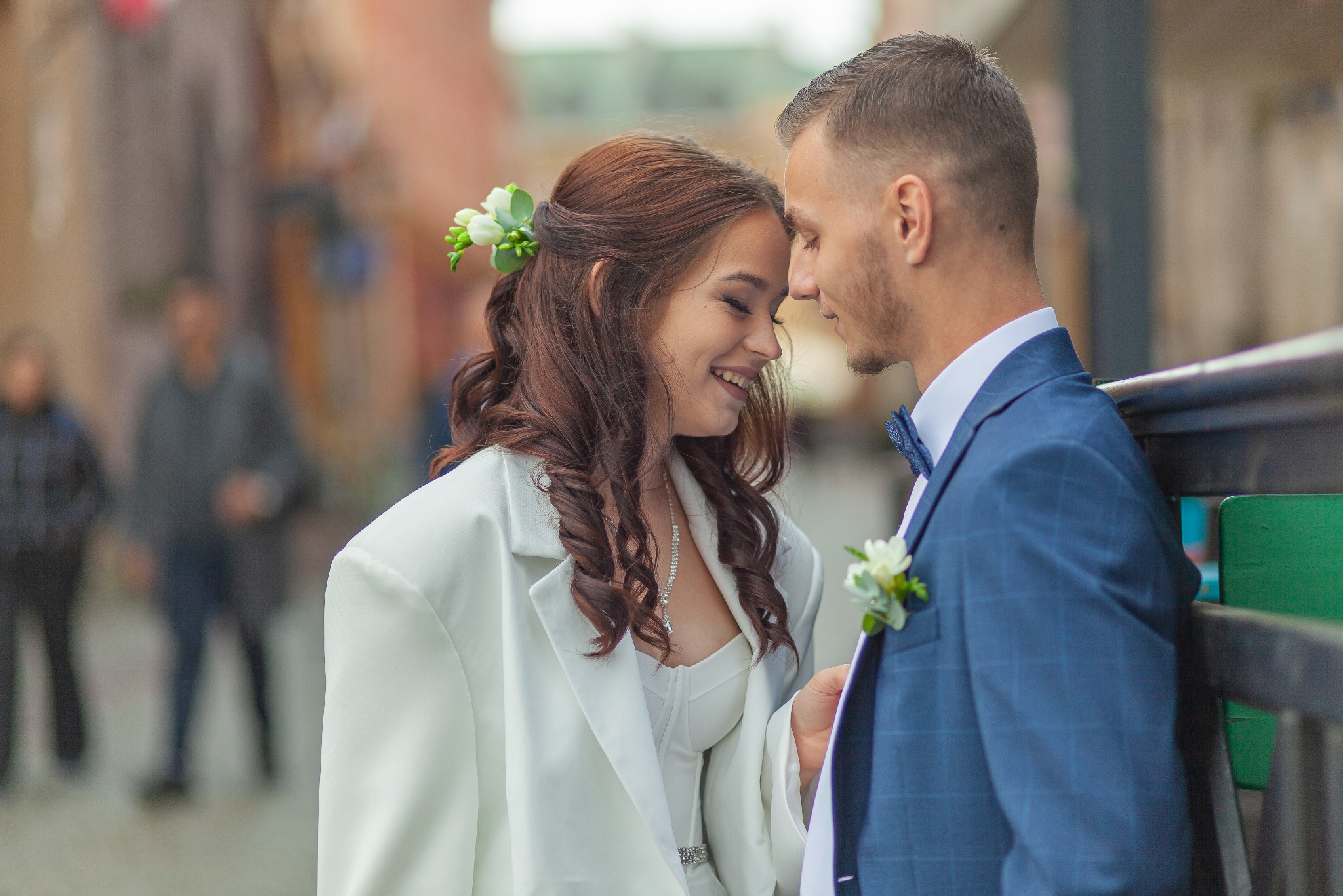 Uśmiechnięta panna młoda z kwiatami we włosach, w białym garniturze, opiera głowę o pana młodego w niebieskim garniturze w kratę z muszką, na tle rozmytego miejskiego krajobrazu.