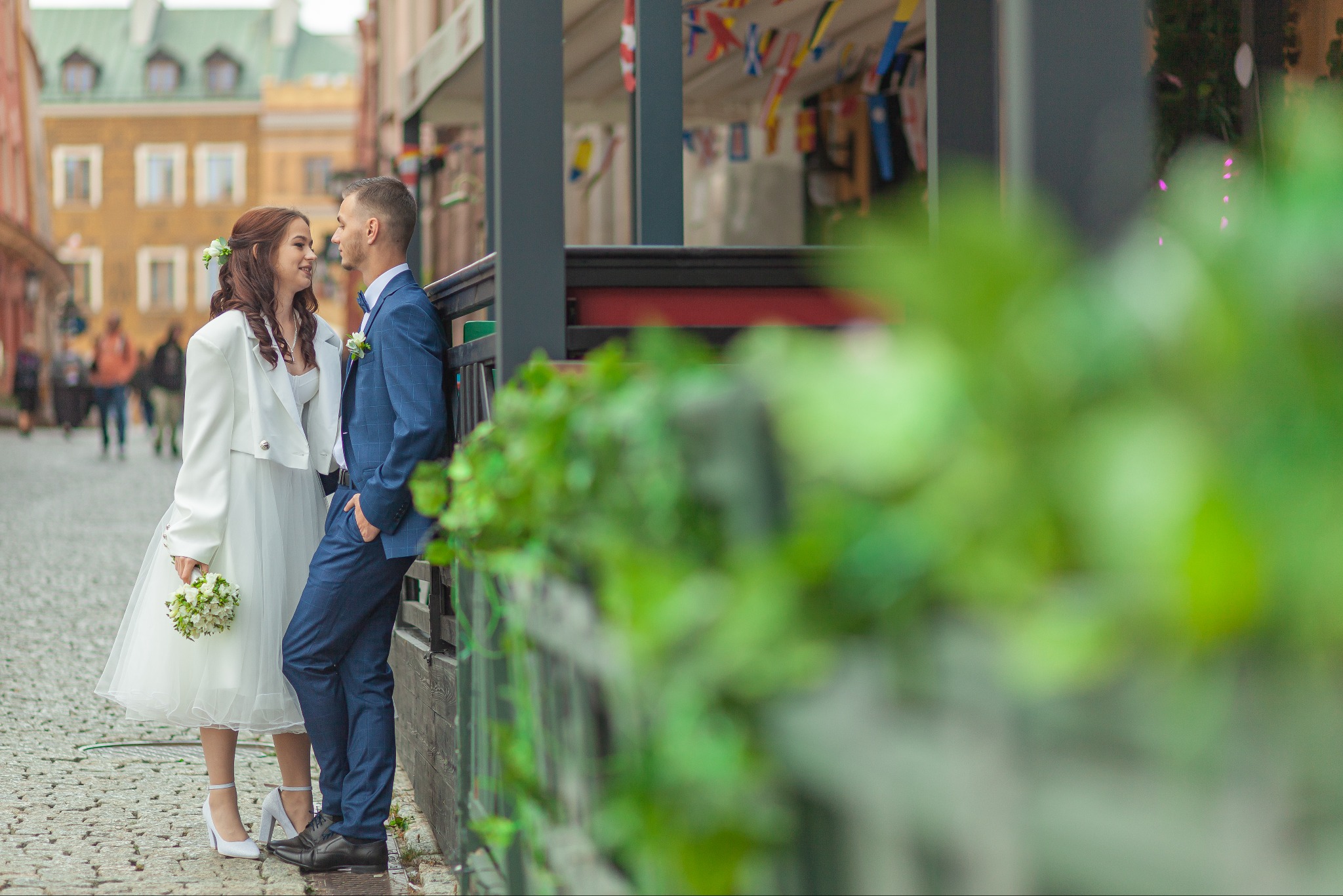 Para młoda w strojach ślubnych, stojąca na tle starego miasta, panna młoda trzyma bukiet, oboje patrzą na siebie z uśmiechem.