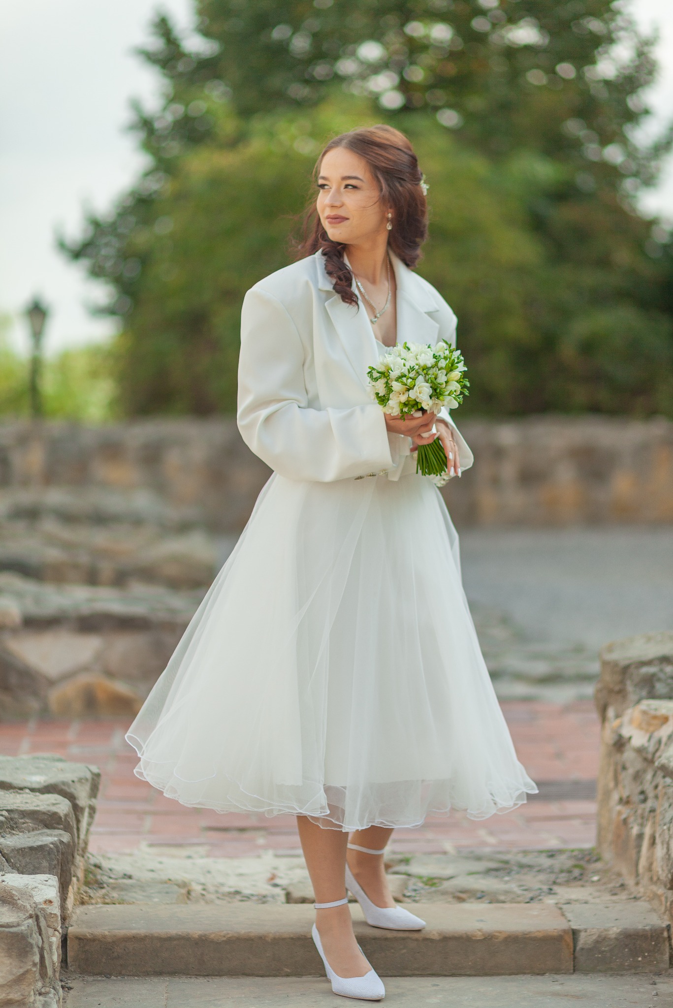 Panna młoda w białej sukience midi i marynarce oversize, trzymająca bukiet białych frezji, z delikatnym makijażem i upiętymi włosami, pozuje na tle kamiennych schodów i zieleni.