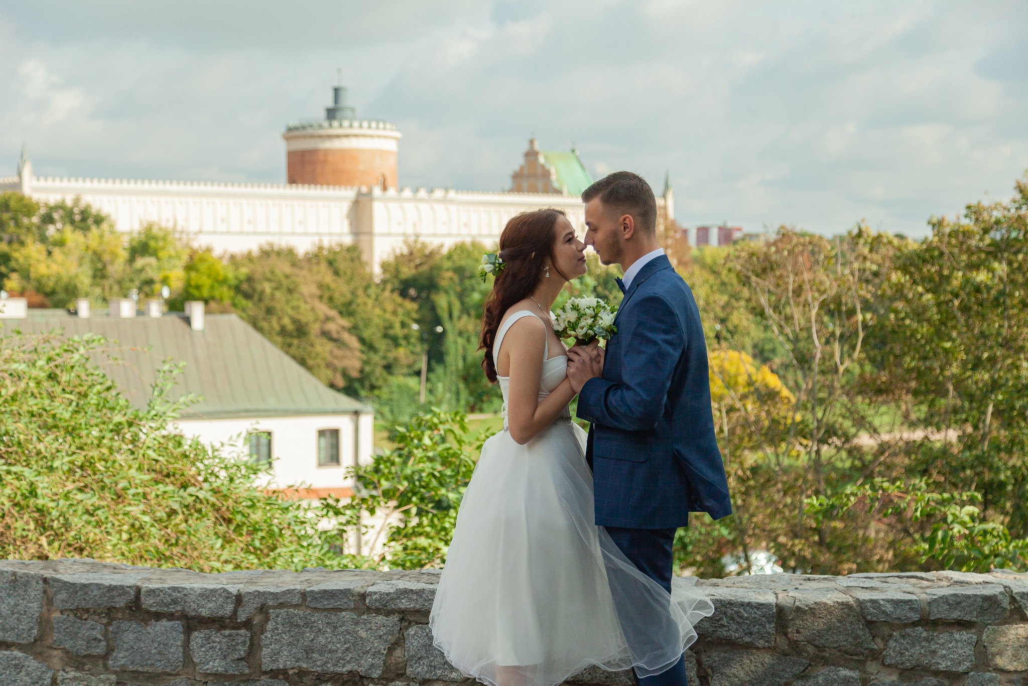 Para młoda w objęciach na tle Zamku w Lublinie, panna młoda z bukietem białych kwiatów, pan młody w granatowym garniturze z muszką.