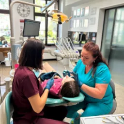 Lekarz stomatolog i asystentka w trakcie badania jamy ustnej pacjentki na fotelu dentystycznym, oświetlonym lampą zabiegową; w tle widoczny zegar i okno.