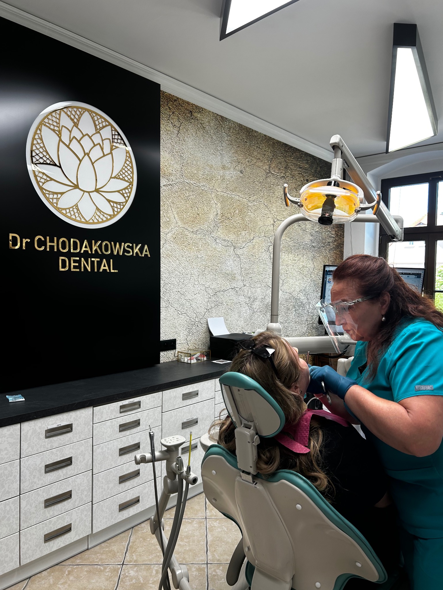 Pacjentka w trakcie zabiegu w gabinecie, lekarz w turkusowym uniformie używa narzędzi, widoczne logo gabinetu Dr Chodakowska Dental z motywem kwiatu lotosu w złotej obwódce na czarnym tle.