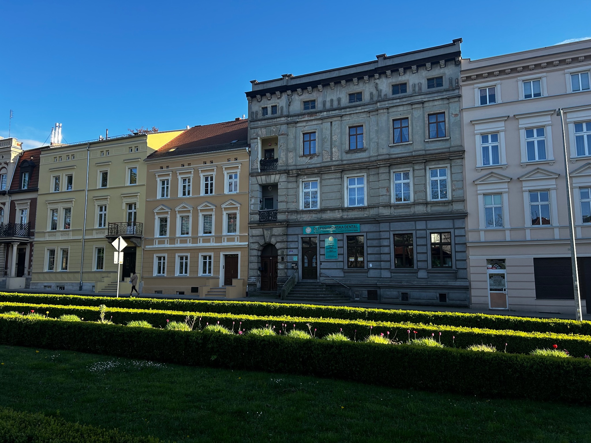 Fasada kamienicy z gabinetem lekarskim 'Dr. Chodakowska Dental' z widocznym wejściem i schodami, otoczona niskim żywopłotem i trawnikiem w słoneczny dzień.