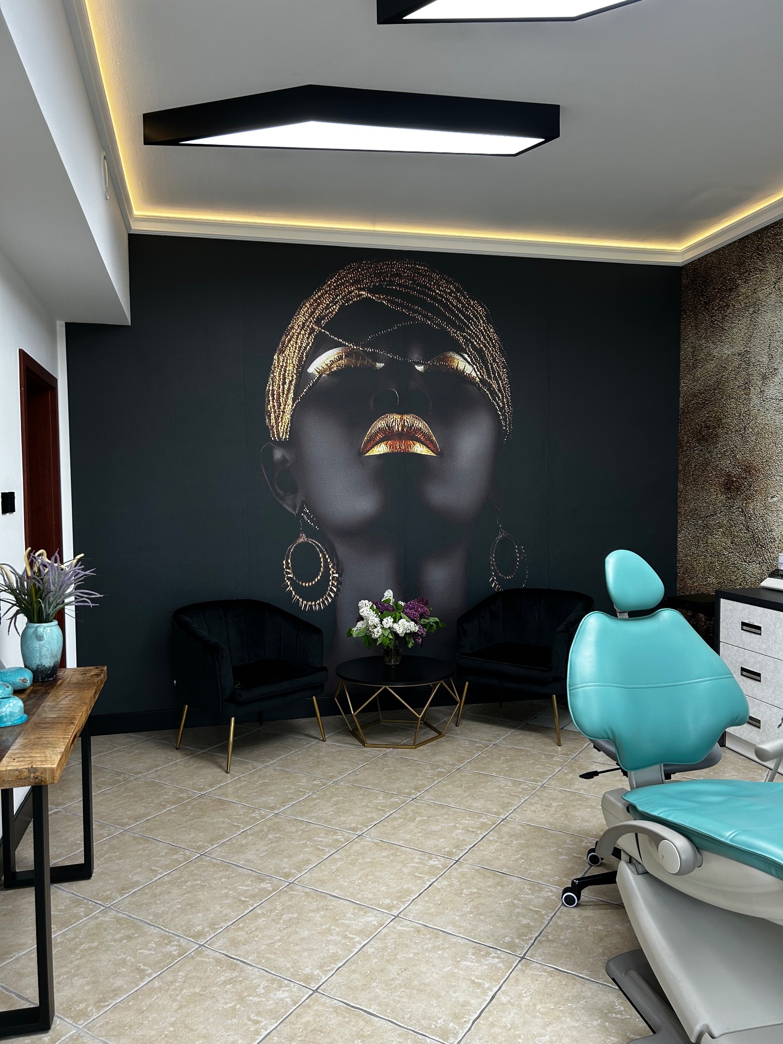 Wnętrze gabinetu z turkusowym fotelem dentystycznym, czarnymi fotelami, stolikiem z kwiatami i dekoracyjną tapetą z portretem kobiety w złotej biżuterii.
