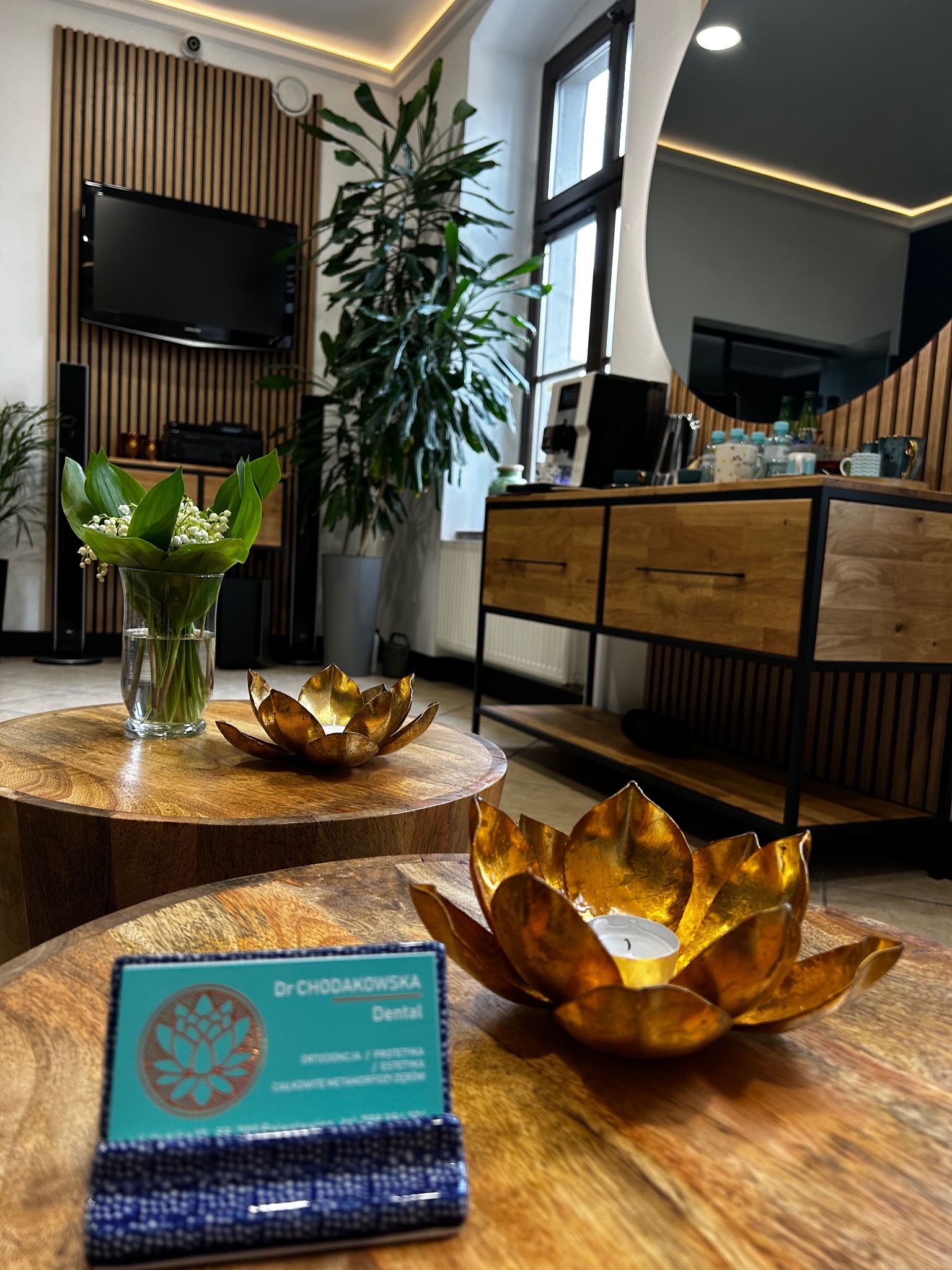 Wnętrze gabinetu stomatologicznego z drewnianymi stolikami, kwiatami w wazonie, świecznikami w kształcie lotosu i wizytówką Dr Chodakowskiej Dental.