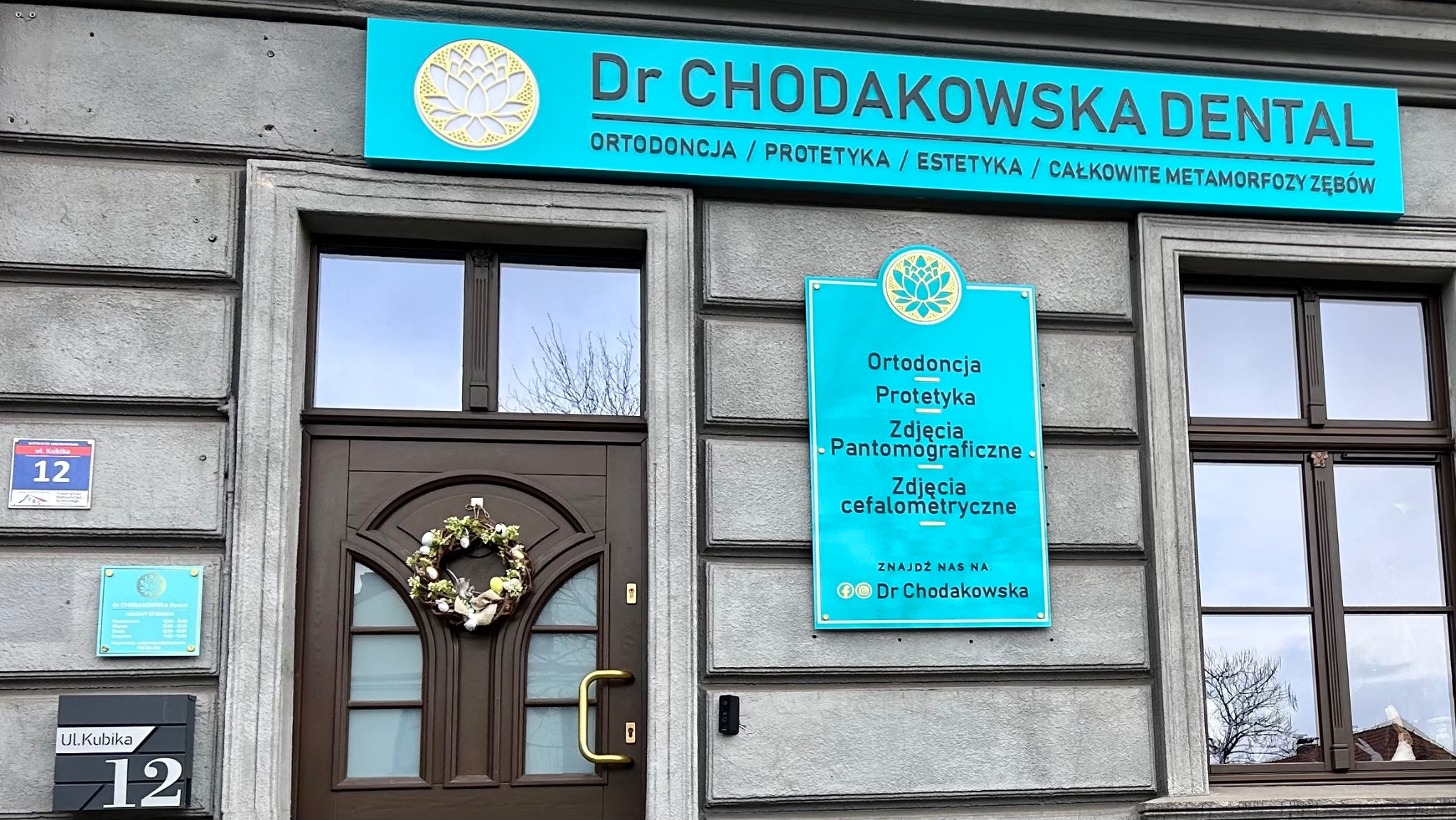 Fasada budynku z szyldem 'Dr Chodakowska Dental' oferującego ortodoncję, protetykę, estetykę i całkowite metamorfozy zębów, z dekoracyjnym wieńcem na drzwiach wejściowych.